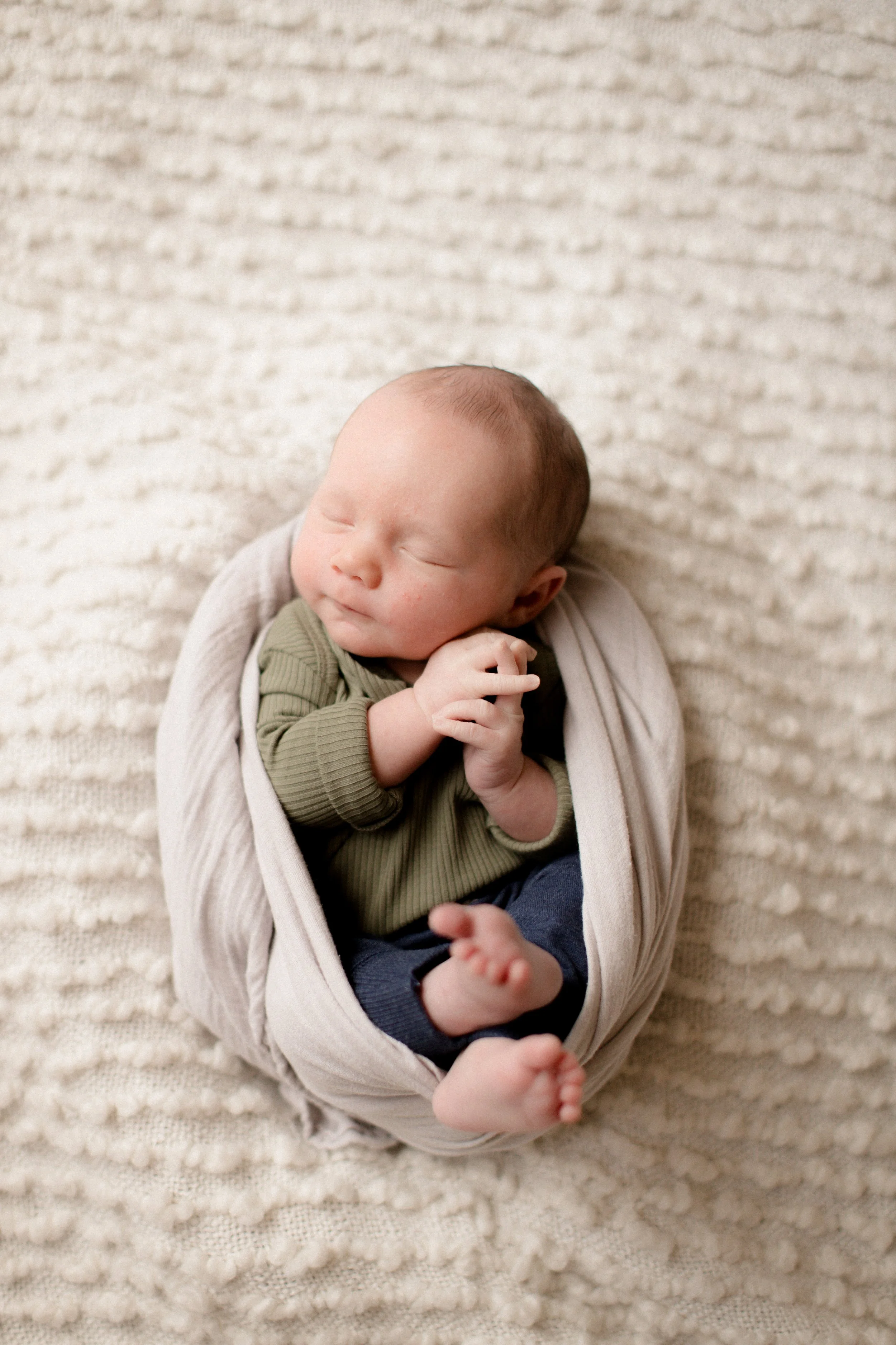 A sleeping baby wrapped in a white blanket lying on a textured beige rug. Lifestyle newborn session shot in Chicago IL.