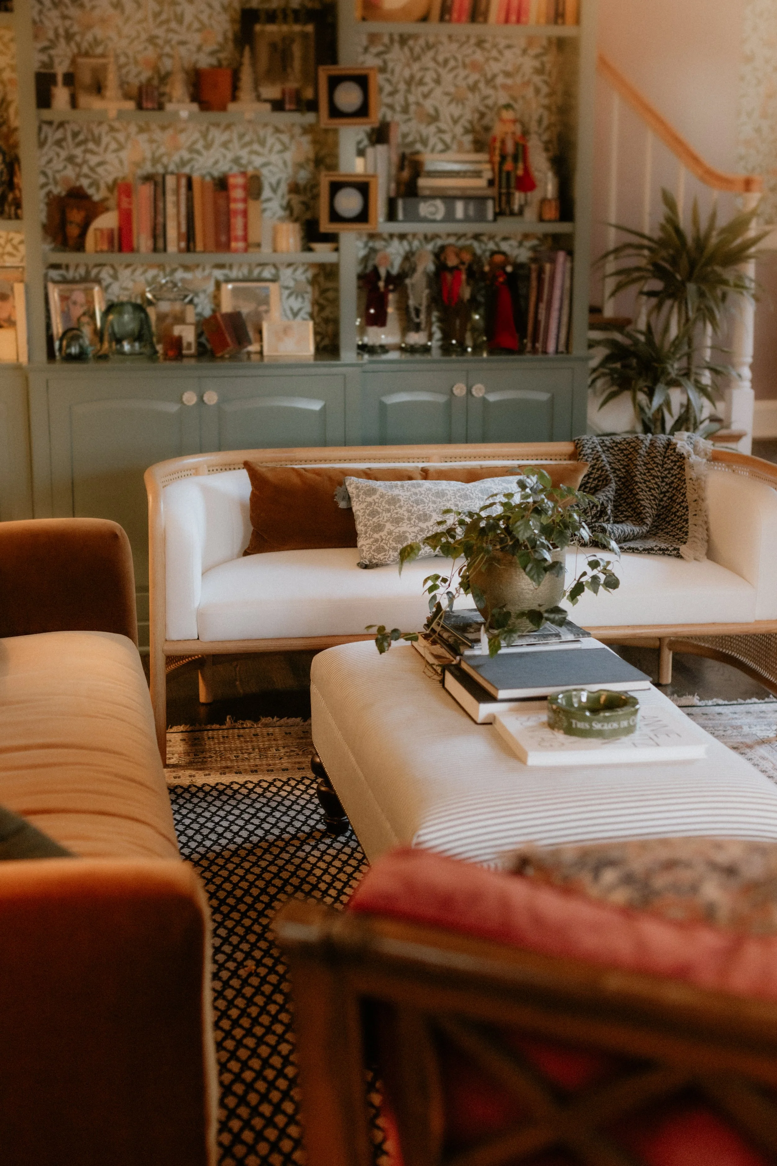 Cozy living room with a white sofa, brown and patterned pillows, a coffee table with books and a green plant, and a green bookshelf with various decorative items, books, and framed photos, against floral wallpaper and a large potted plant. 