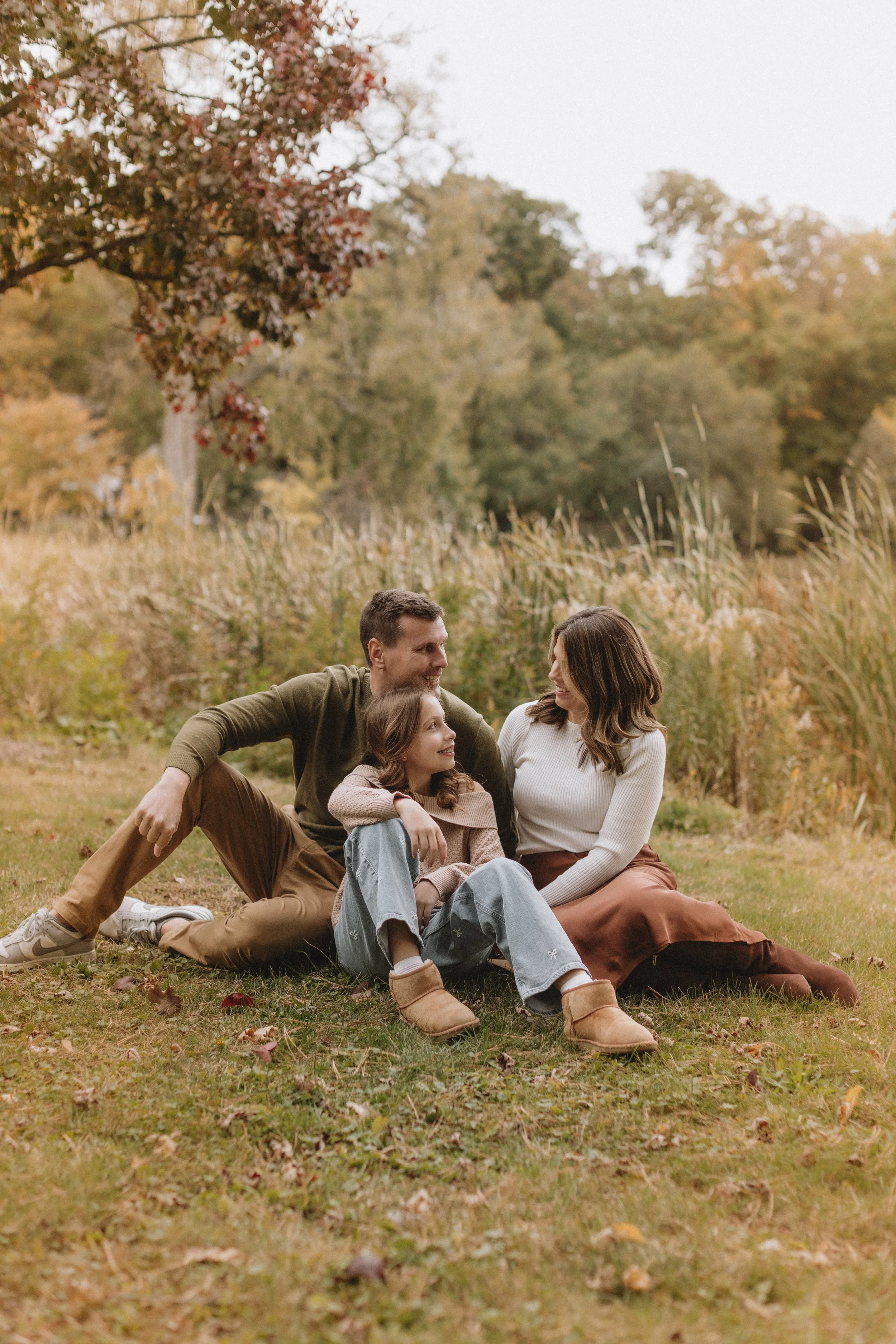 A family of three sitting on the grass in an autumn park, smiling and looking at each other, with trees and tall grasses in the background. Family photography session Evanston IL.  
