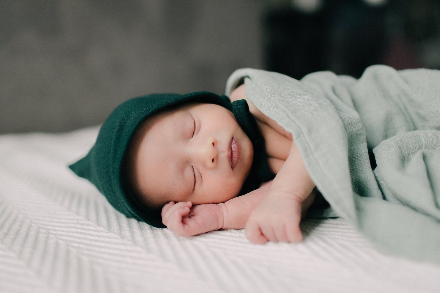 A sleeping baby in a green hoodie, lying on a white textured blanket with one hand near their face. Newborn photography in Chicago IL