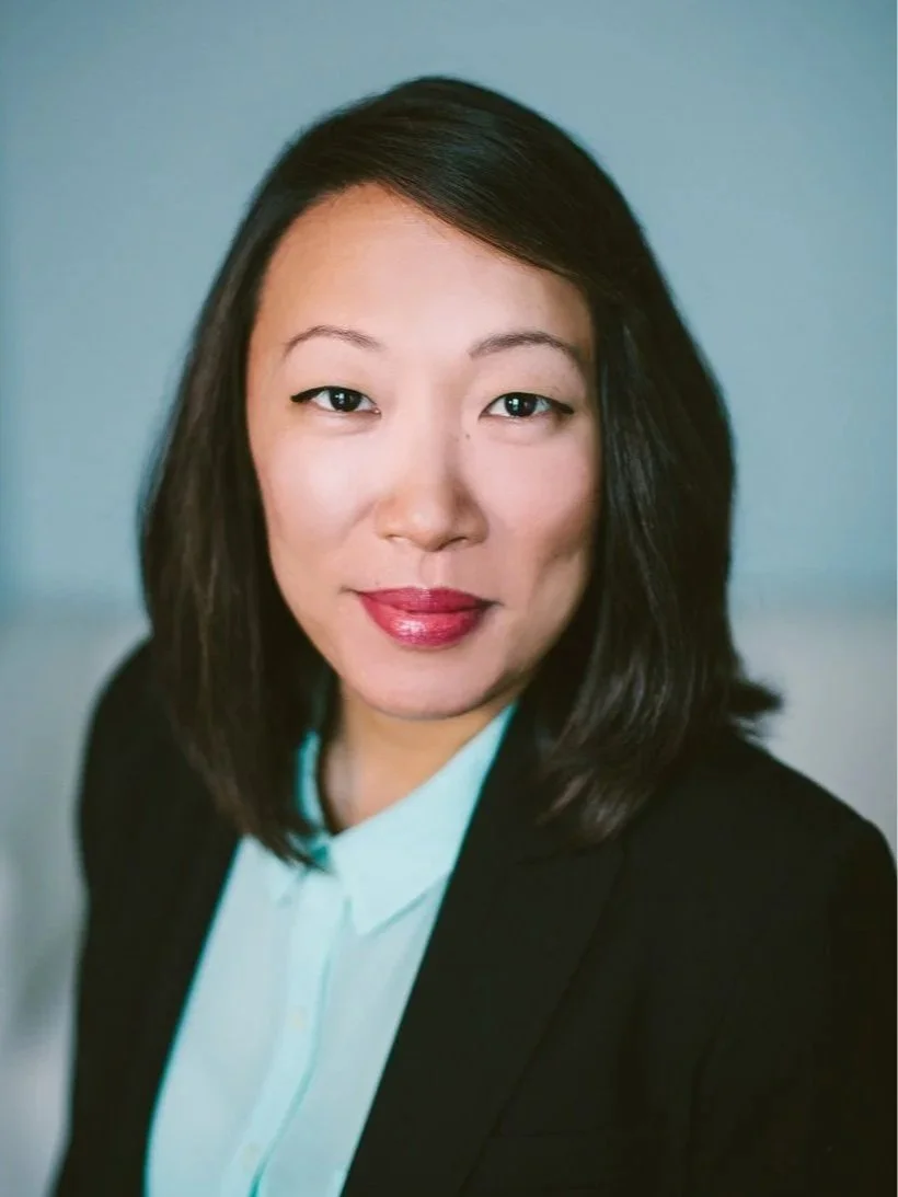 A professional headshot of an Asian woman with shoulder-length dark hair, wearing a black blazer and a white blouse, smiling slightly against a plain blue background.