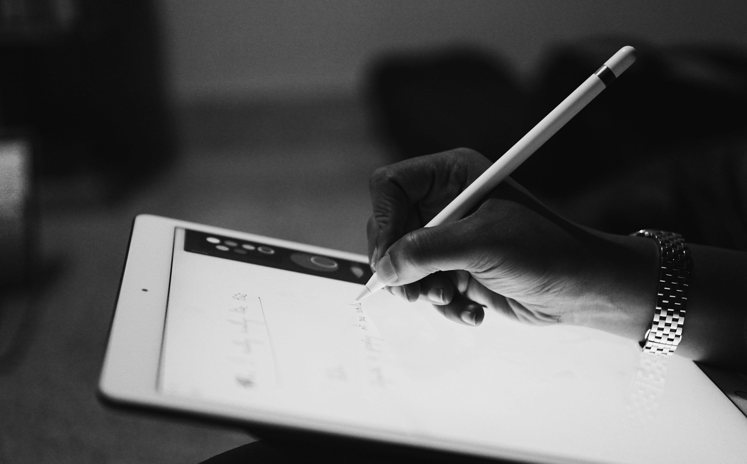 A close-up of a hand holding a stylus pen and writing on an electronic tablet or e-reader with a black background.