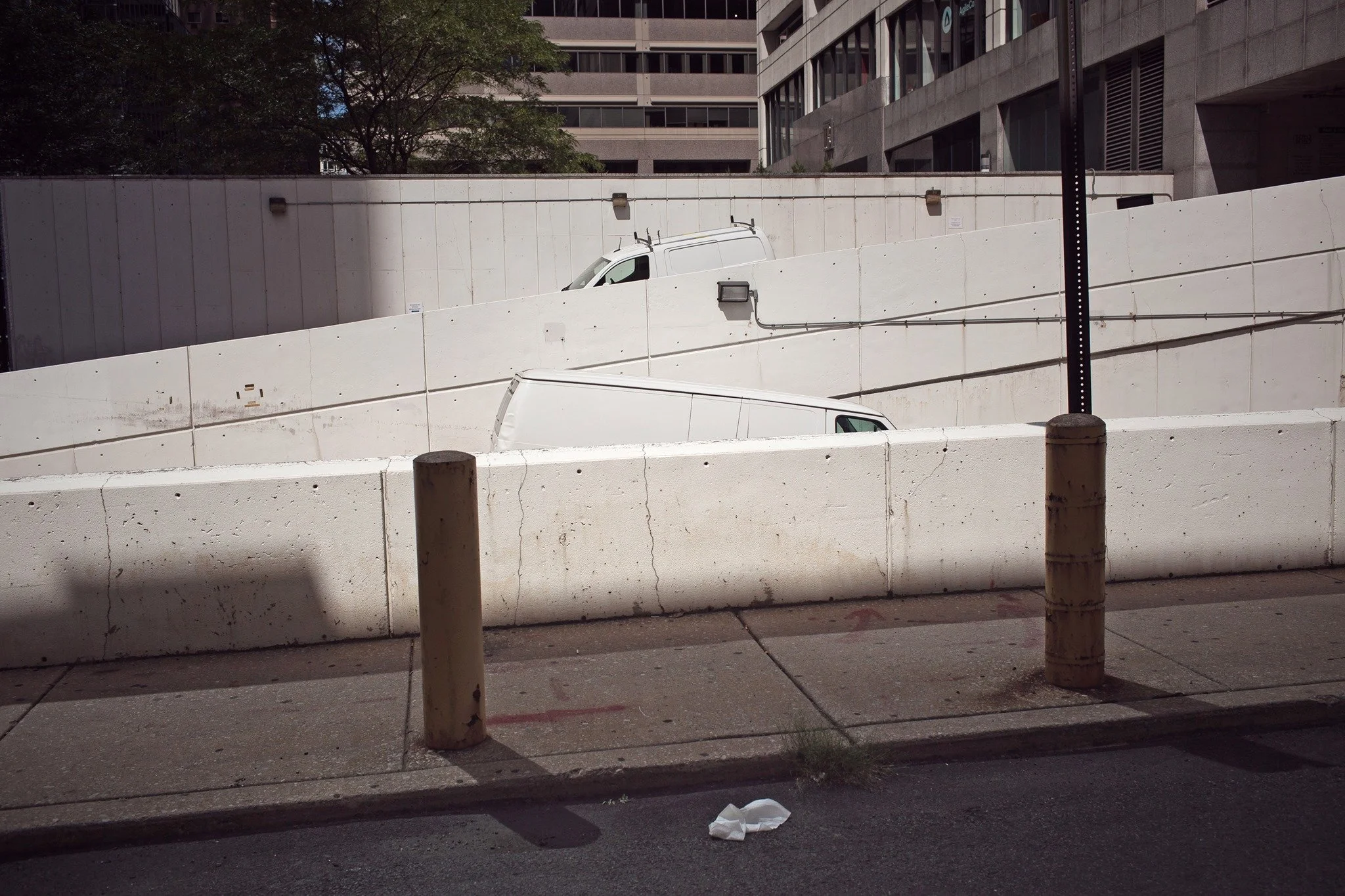 Concrete barriers on a city sidewalk, with a white van partially visible behind the barriers, and a street lamp post in the foreground. Behind that, a building with multiple windows and trees are visible.