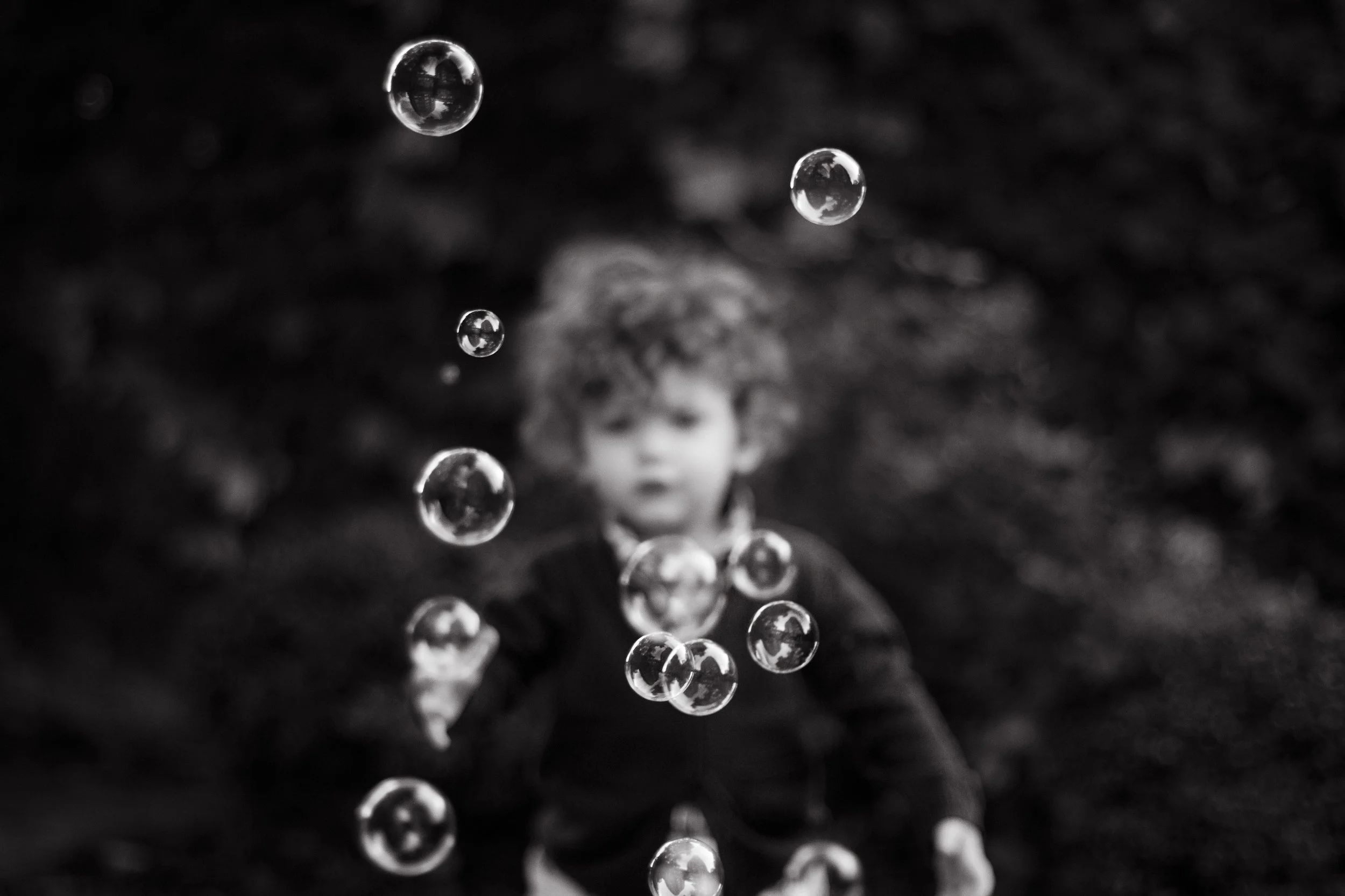 A young boy with curly hair playing with bubbles outdoors, with a blurred natural background. Black and white photography with an editorial style.  Evanston and Chicago.