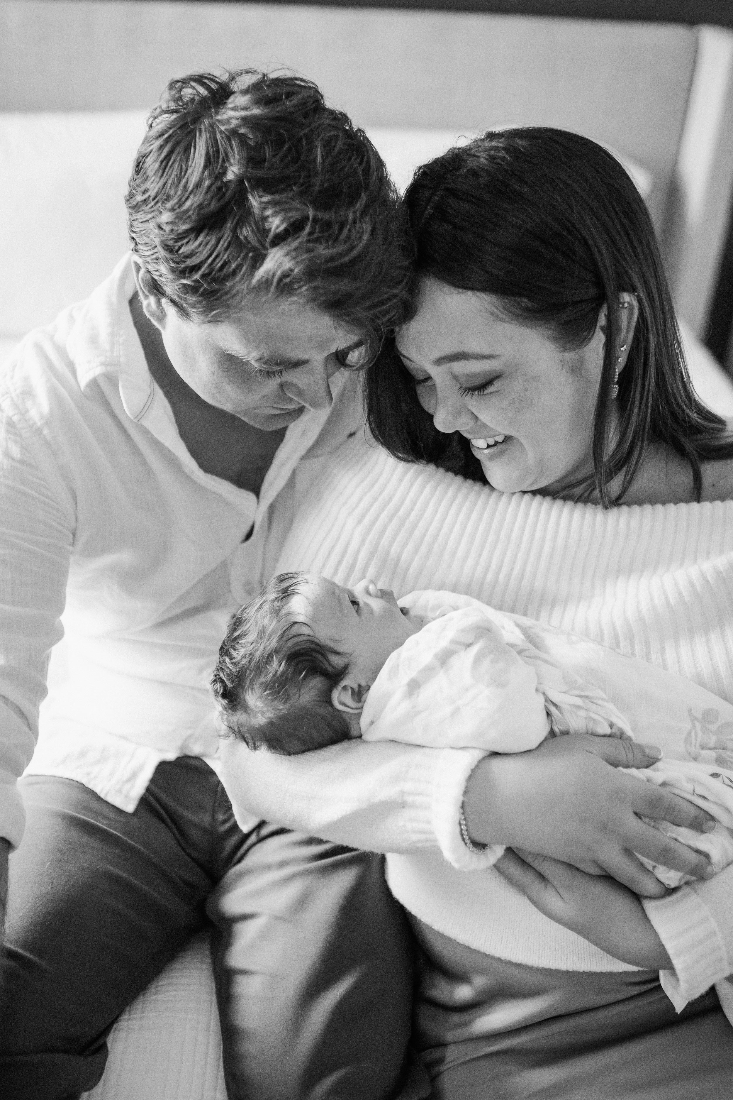A black and white photo of a family with a mother, father, and baby. The parents are leaning close to the baby, who is lying in the mother's arms, with all three smiling and showing affection. Lifestyle newborn session shot in Evanston IL.