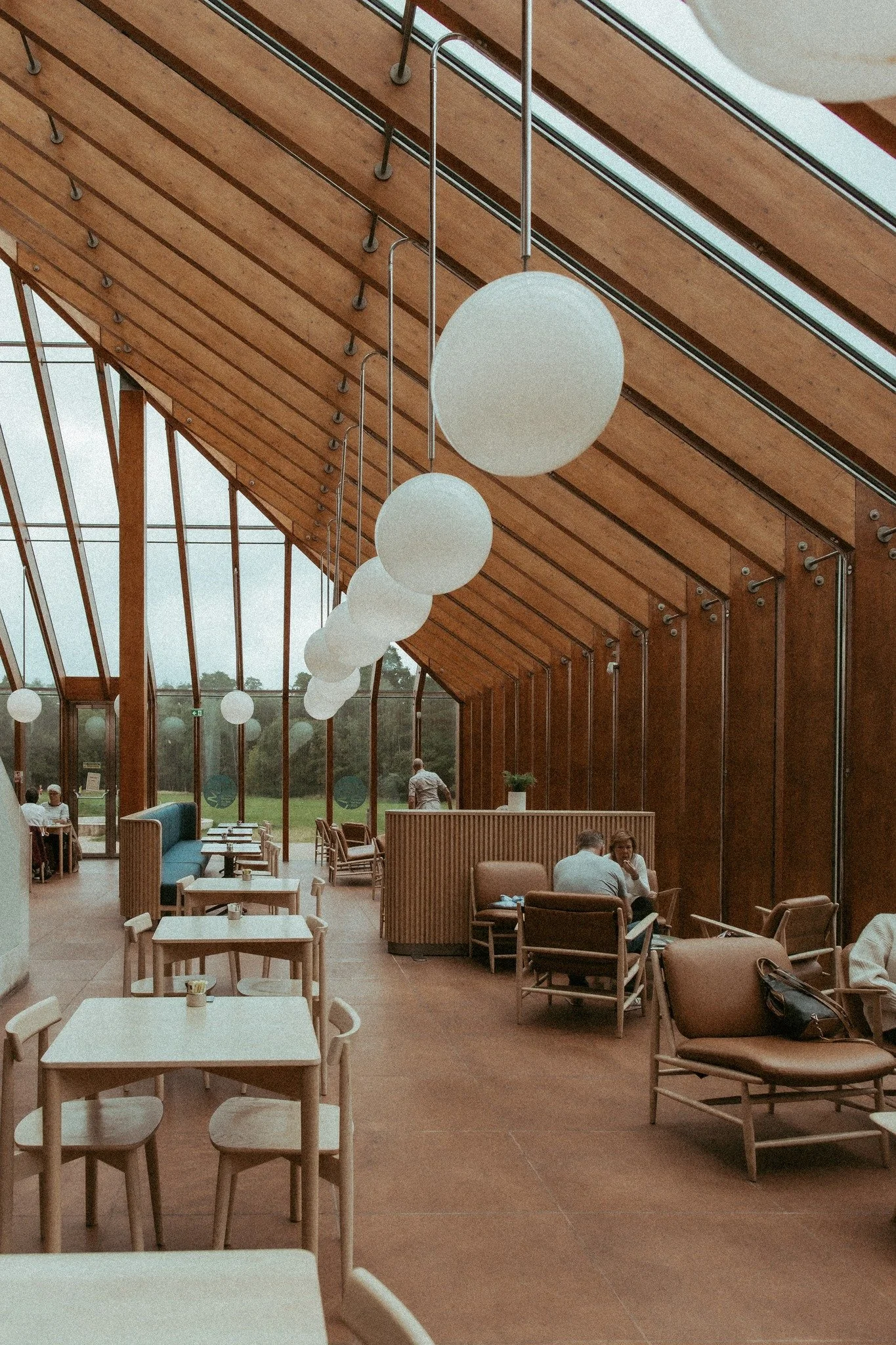 Interior of a modern café with wooden walls, hanging spherical pendant lights, and large windows revealing a green outdoor scene. Interior Design and Architectural Photography Chicago IL, Erin Drewitz Photography