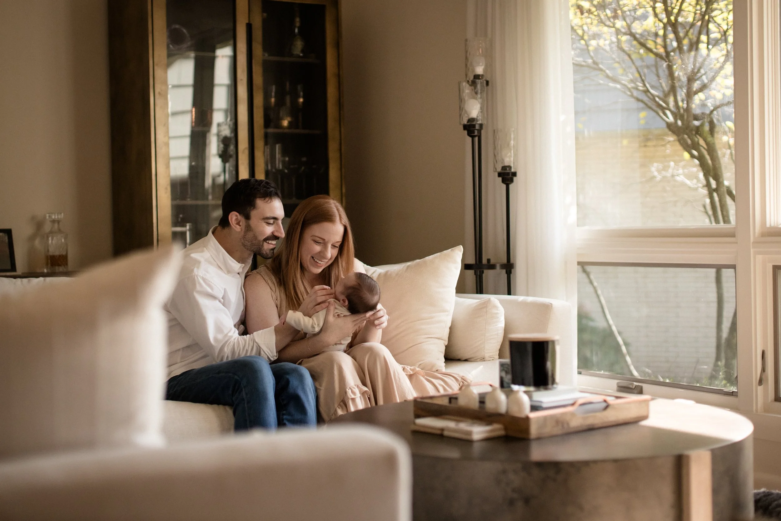 A happy family sitting on a beige sofa in a living room, with a woman holding a baby and a man smiling at the baby. Sunlight streams through a large window with sheer curtains, illuminating the room. Lifestyle newborn session shot in Evanston IL.