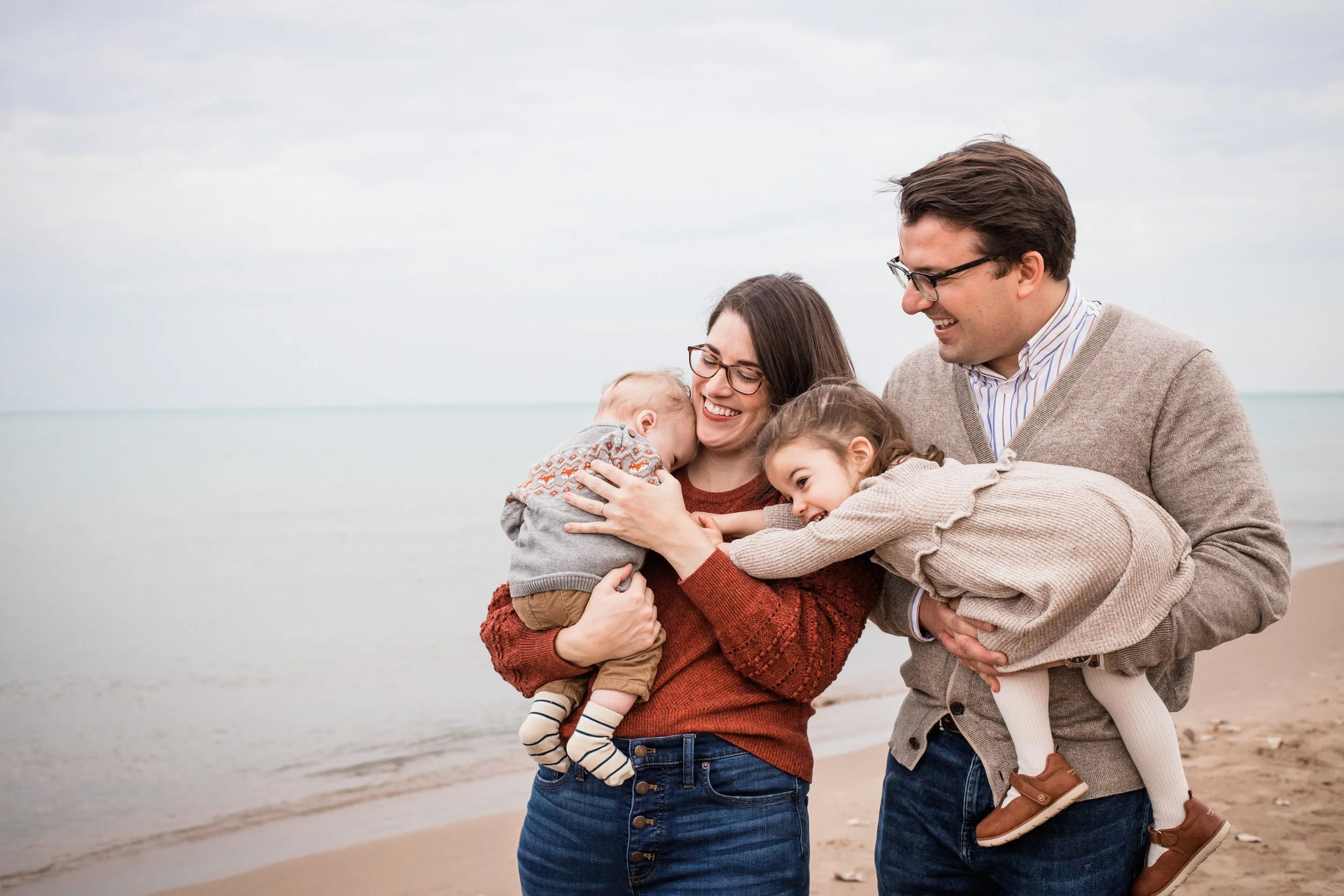 A happy family of four, including a mother, father, and two young children, hugging and playing on the beach near the water.
