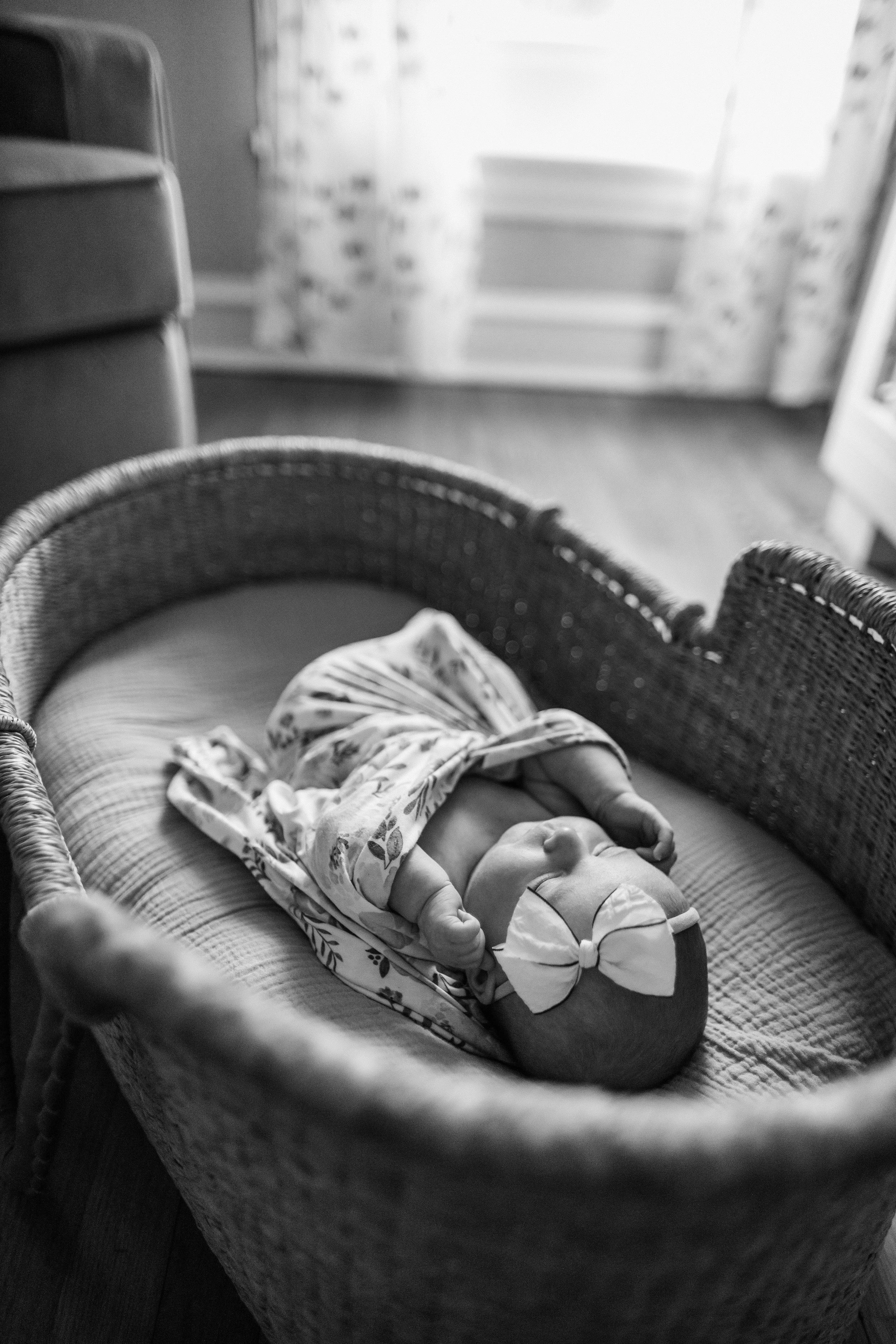 A newborn baby wearing a headband with a bow, lying in a wicker crib indoors.