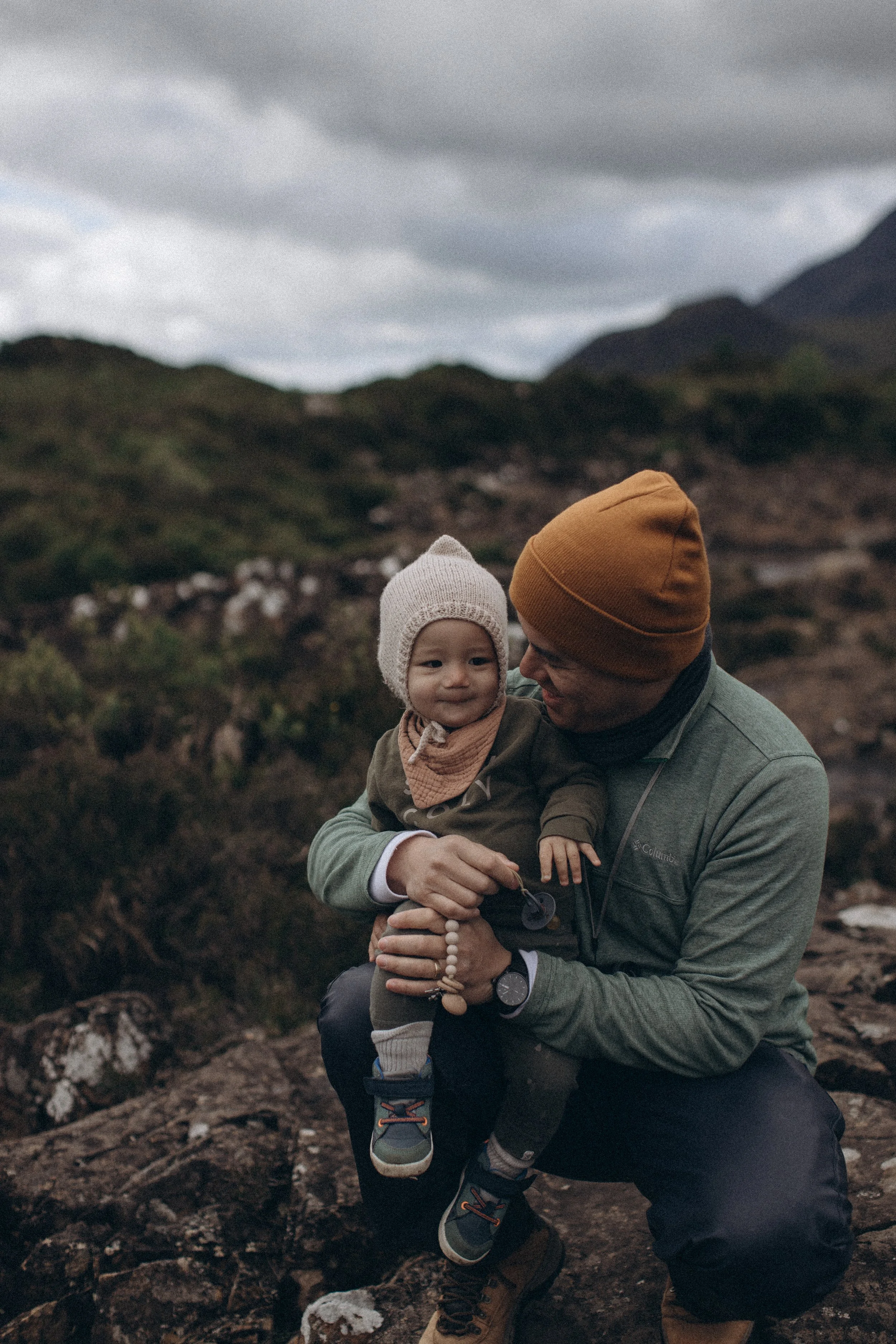 A man holding a smiling toddler outdoors on a rocky terrain with mountains and cloudy sky in the background.