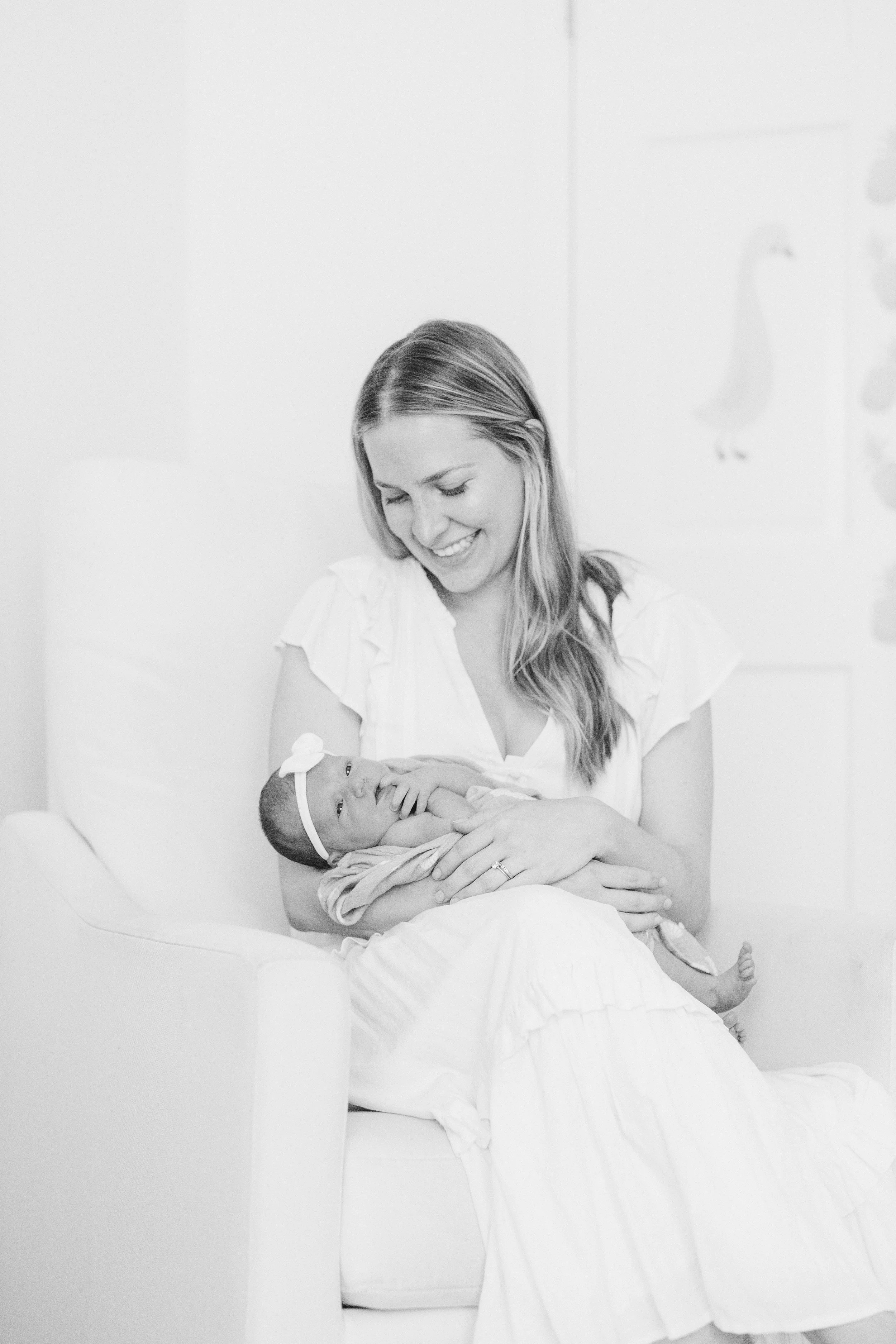 A woman sitting in a chair holding a newborn baby girl, both smiling and looking at each other, in a softly lit room. Lifestyle newborn session shot in Chicago IL.