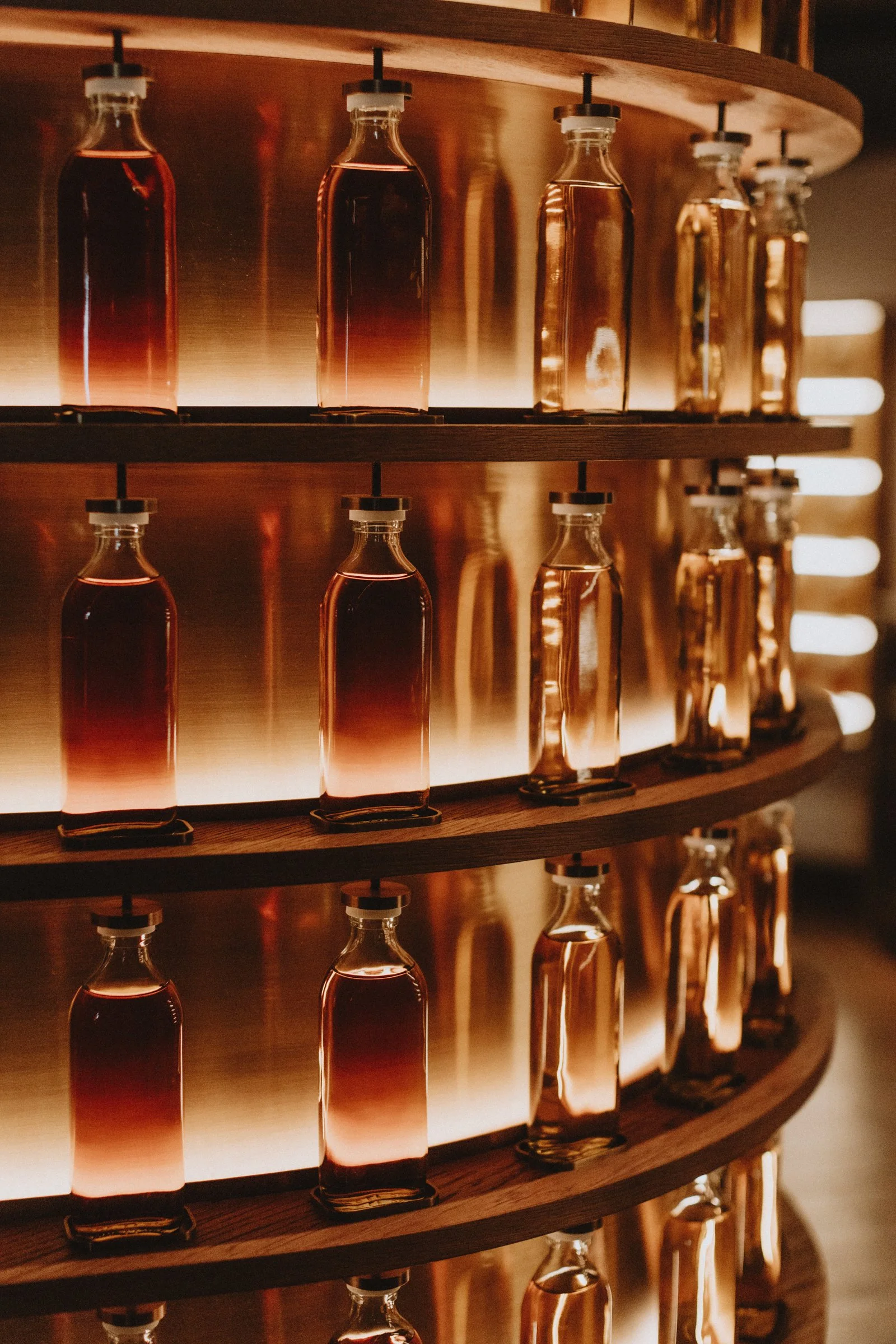Glass bottles filled with amber-colored liquid, displayed on curved wooden shelves with backlighting. Product Photography Chicago IL, Erin Drewitz Photography