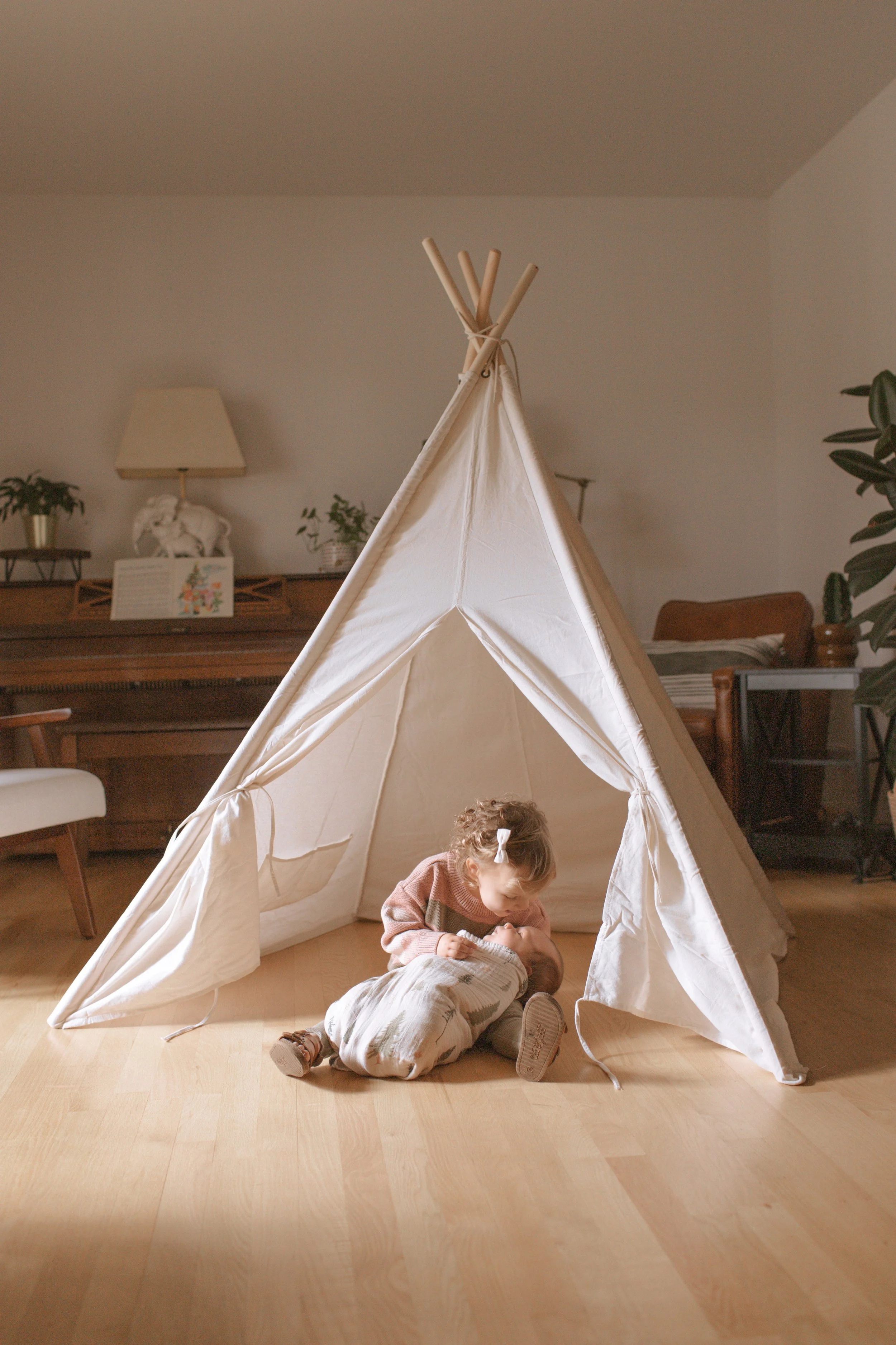 A young girl in a pink sweater is playing with a baby inside a beige fabric teepee tent on a wooden floor in a cozy living room.
