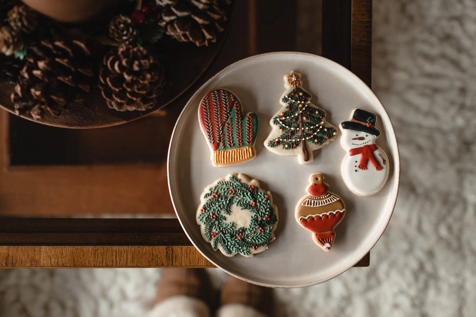 Christmas-themed decorated cookies on a plate, including a mitten, a Christmas tree, a snowman, a holly wreath, and a Christmas ornament. Pine cones are in the background. Product Photography Shoot Evanston, IL.