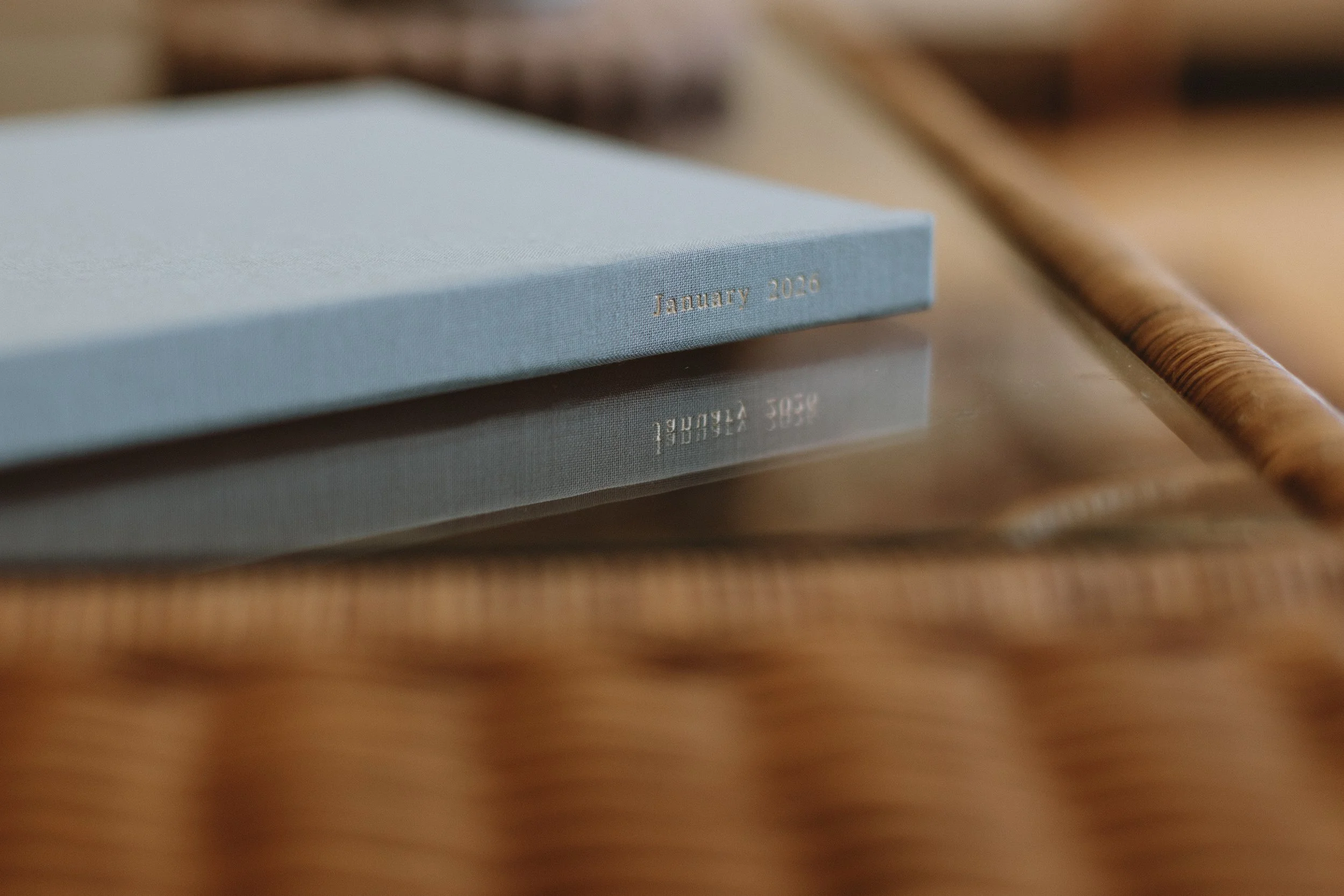 A light blue hardcover journal with 'January 2020' embossed in gold on the bottom right corner of the cover. The journal is resting on a wooden surface, with a wooden rolling pin partially visible beside it. The image also shows a reflection of the journal on the shiny surface beneath it.