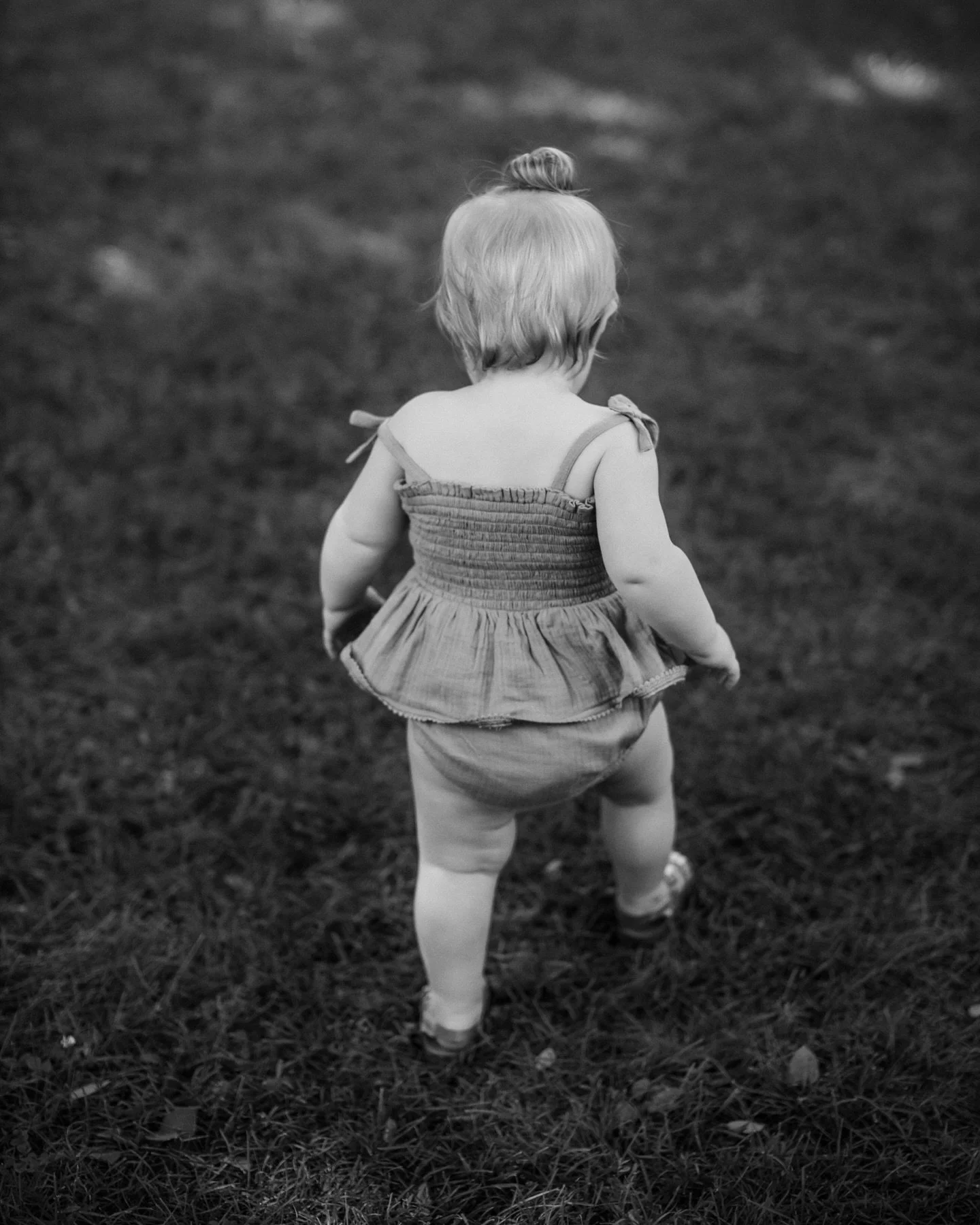 Back view of a young child with short hair, wearing a dress and shorts, standing on a grassy field.