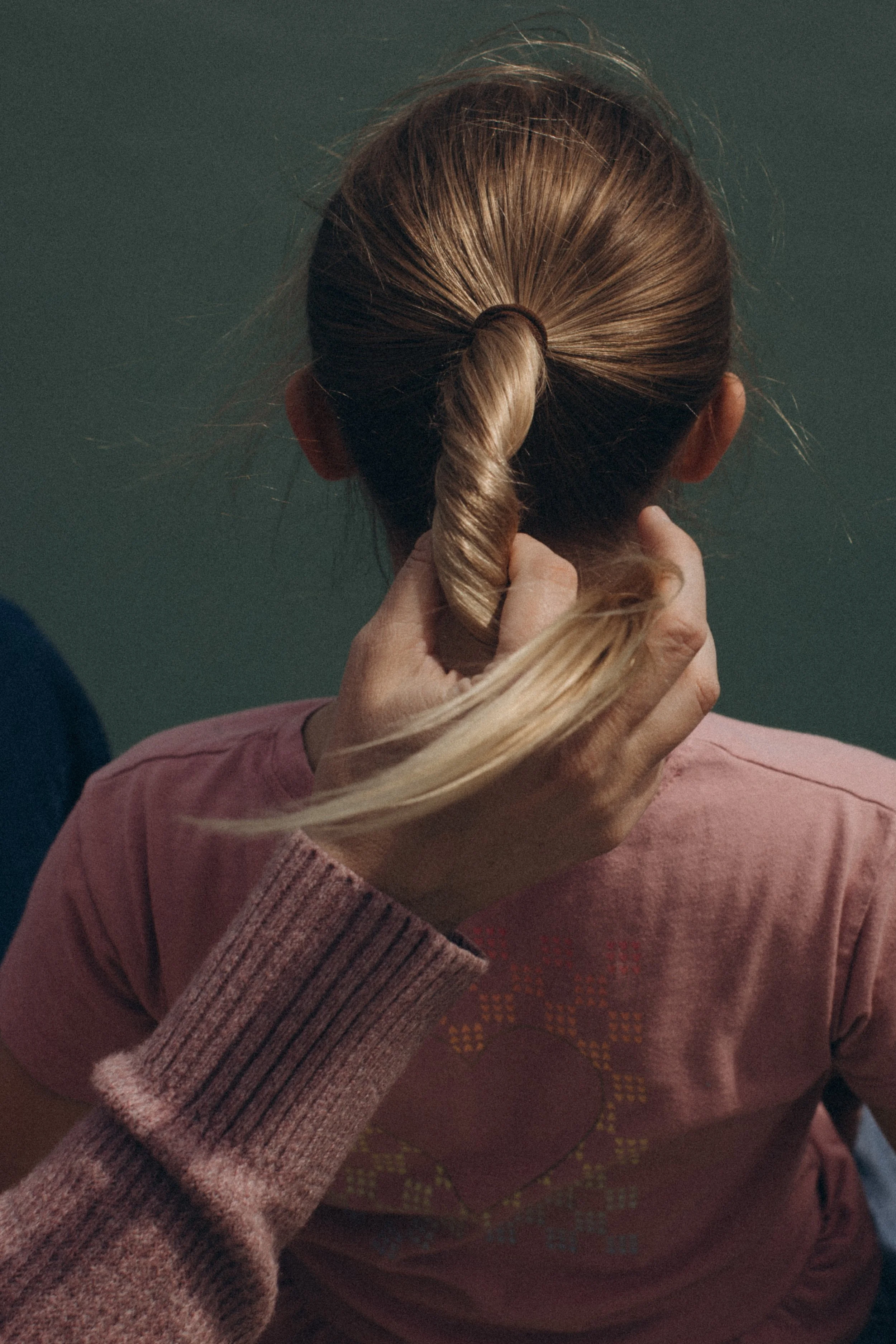 A person with blonde hair tied in a bun, wearing a pink sweater, is holding and twisting their hair in front of their head. The background appears to be a plain wall.