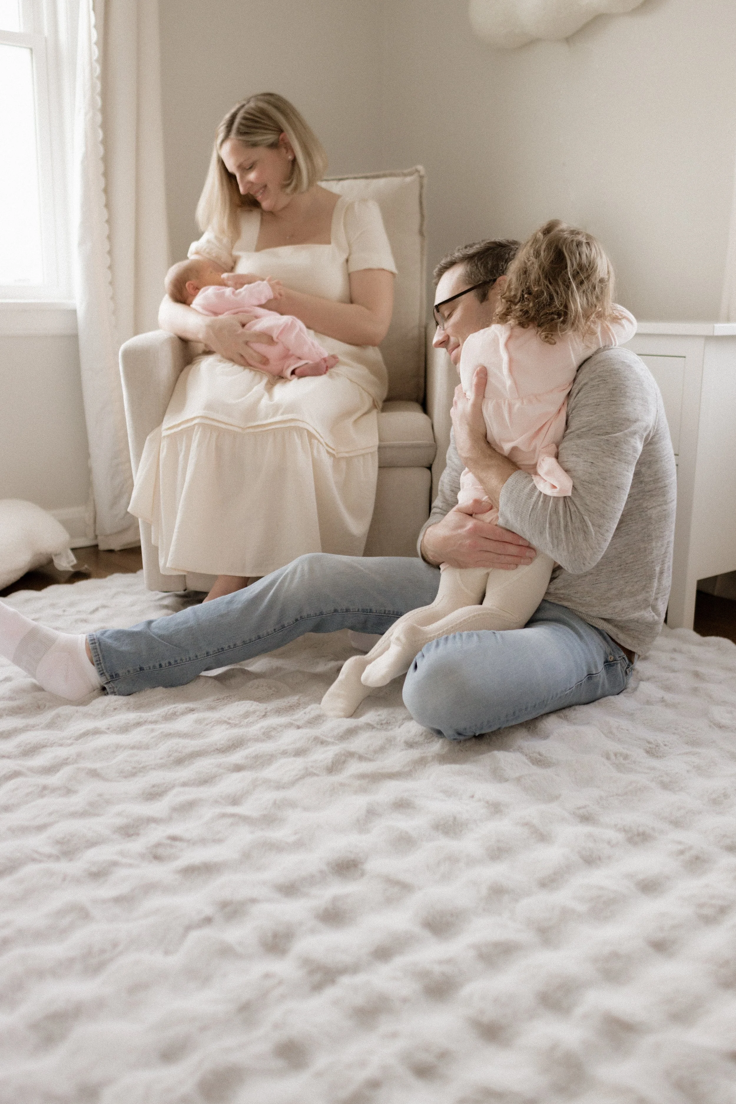 A family of four, including a woman, a man, a girl, and a newborn baby, sitting on a soft white rug in a cozy bedroom, with the woman holding the baby and the man and girl embracing.