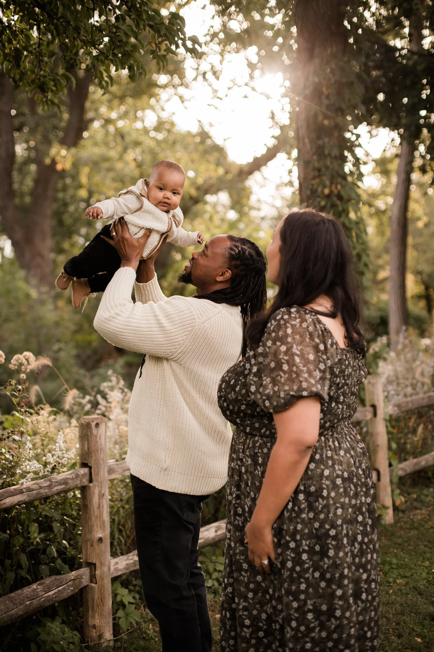 A family outdoors in a park, with a man lifting a baby girl in the air, a woman standing beside them, and trees with sunlight shining through in the background. Gillson Park, Wilmette