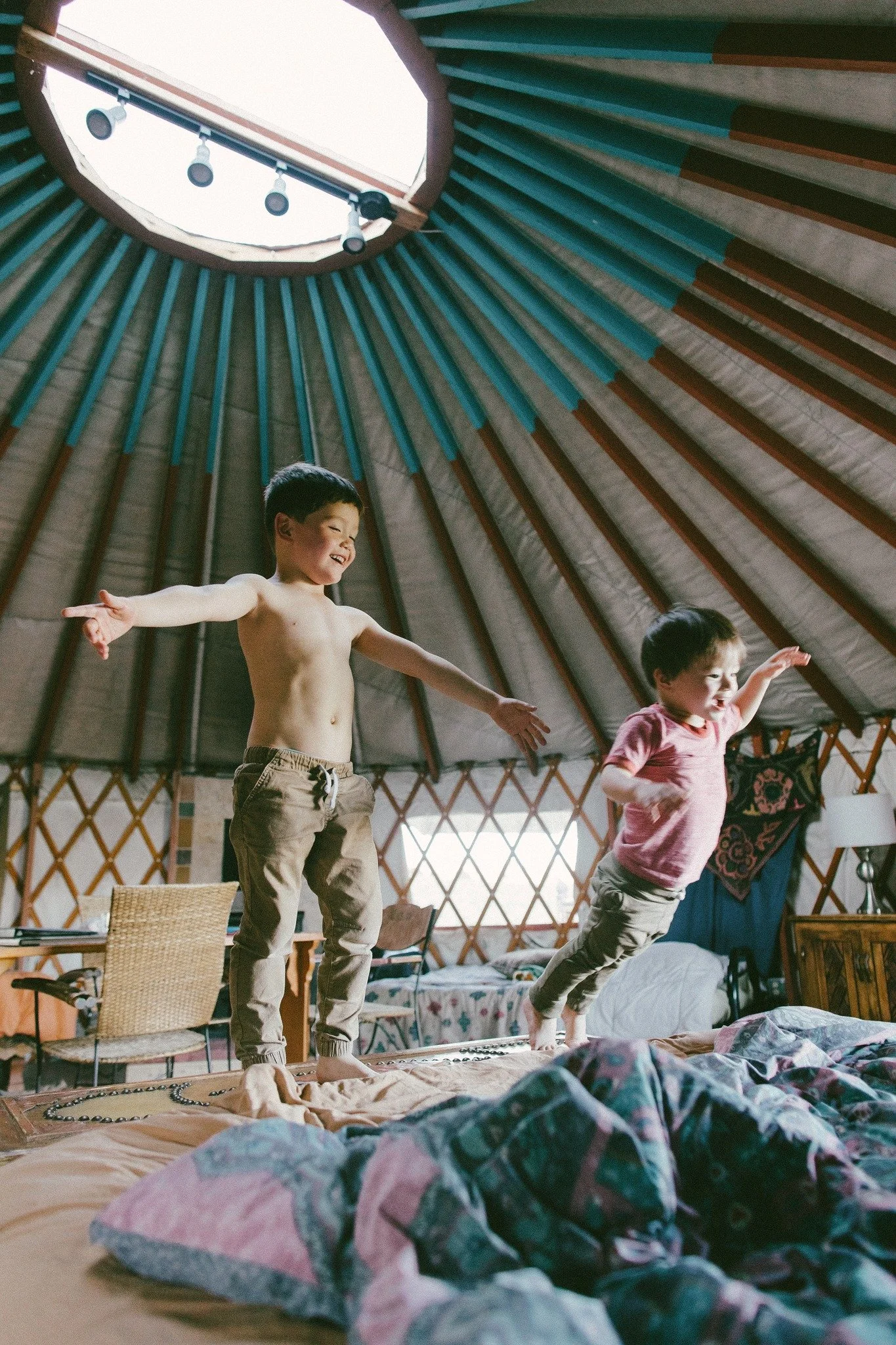 Two children, a boy and a girl, are jumping on a bed inside a yurt-style room with a circular opening at the top.