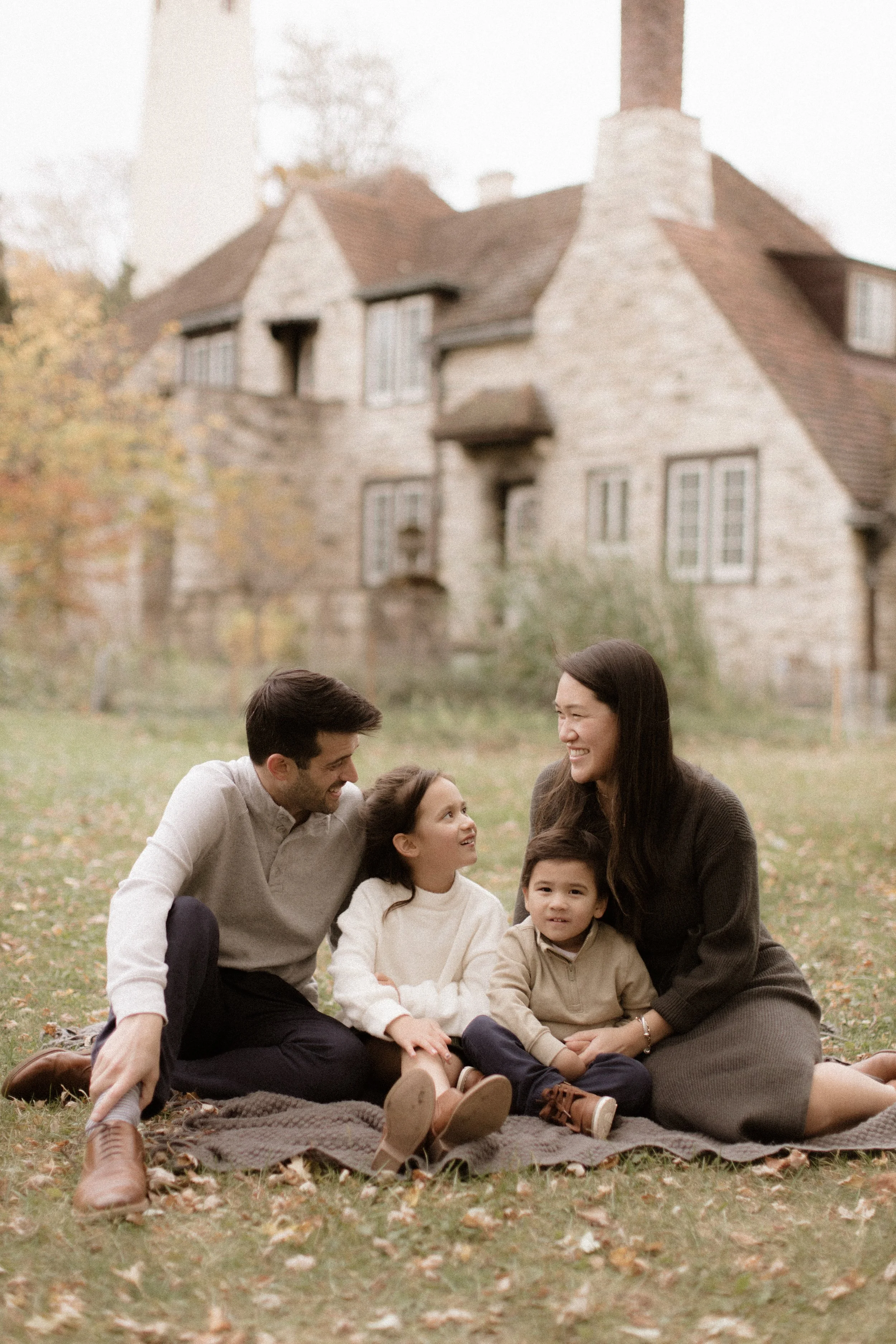 Family of four, including a man, woman, young girl, and young boy, sitting on a blanket on a grassy lawn with a large, old stone house in the background.