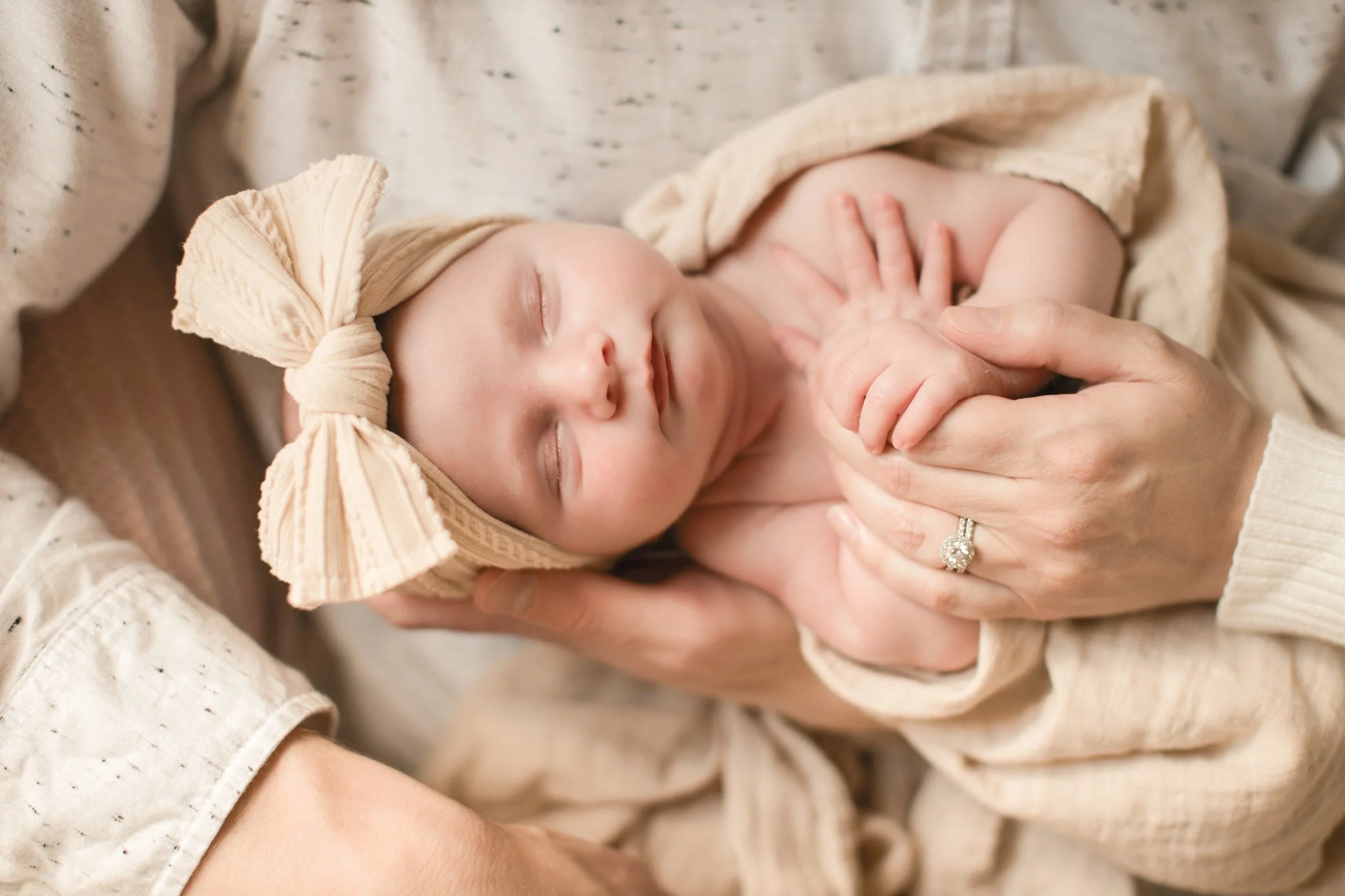 Close-up of a sleeping baby with a beige headband and blanket, being gently cradled by an adult's hands.