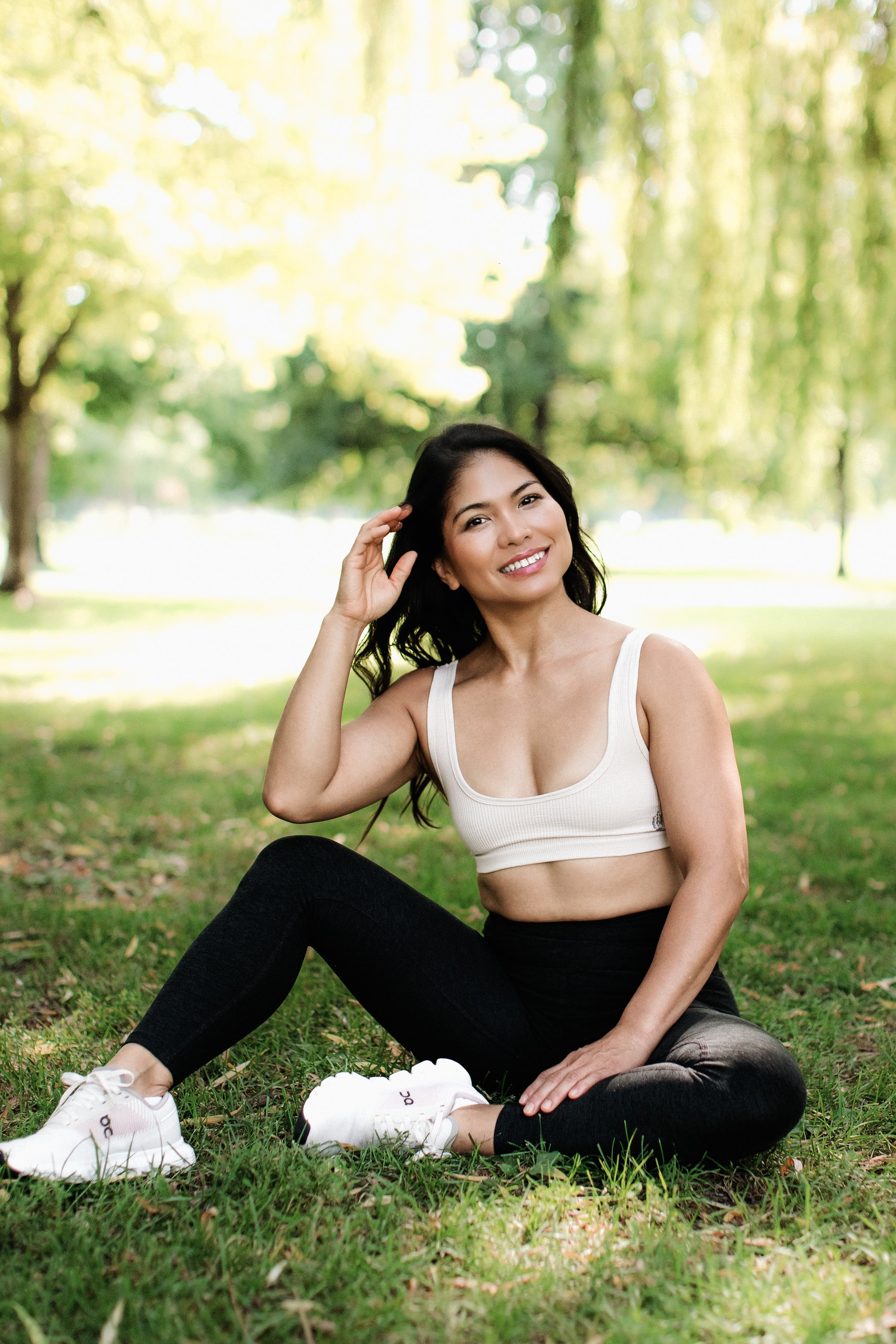 Young woman sitting on grass in a park, smiling, wearing a white sports bra, black leggings, and white sneakers, with trees and sunlight in the background.