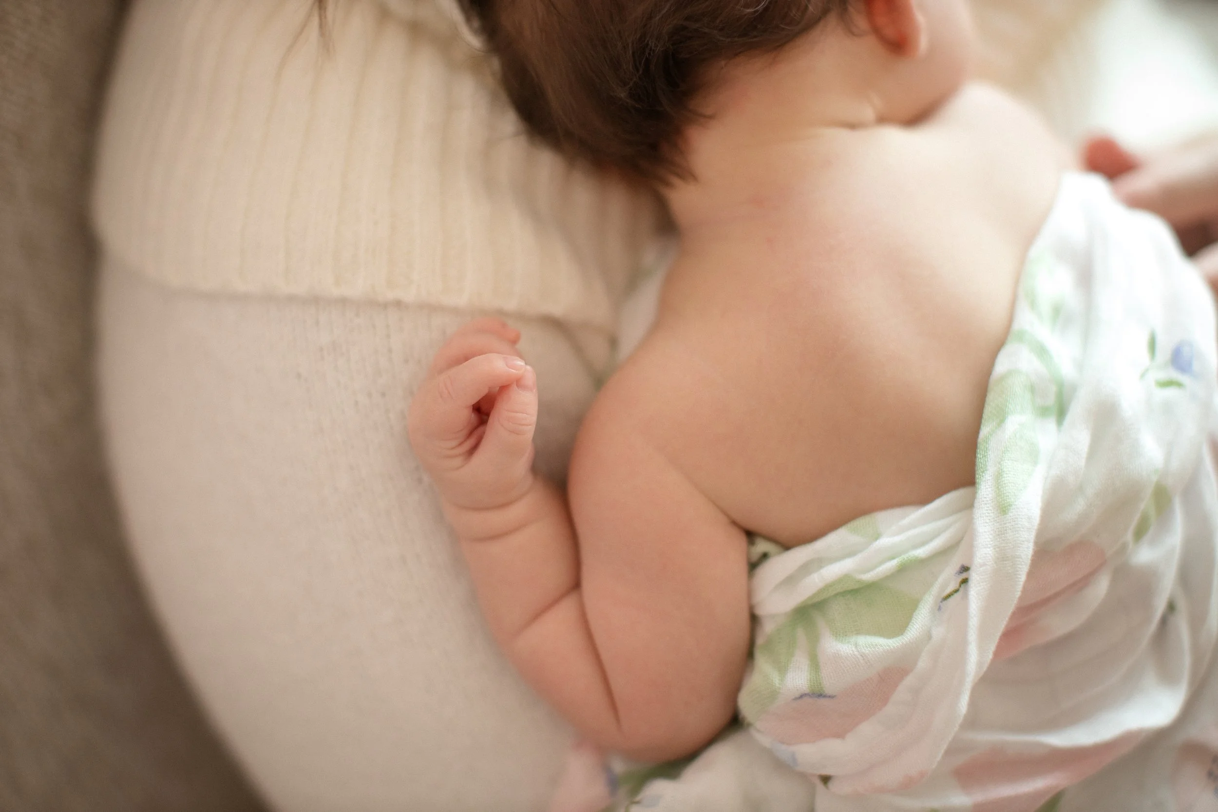 Close-up of a sleeping baby with the baby's head resting on an adult's arm, wearing a white and pastel-colored blanket. Lifestyle newborn session shot in Chicago IL.