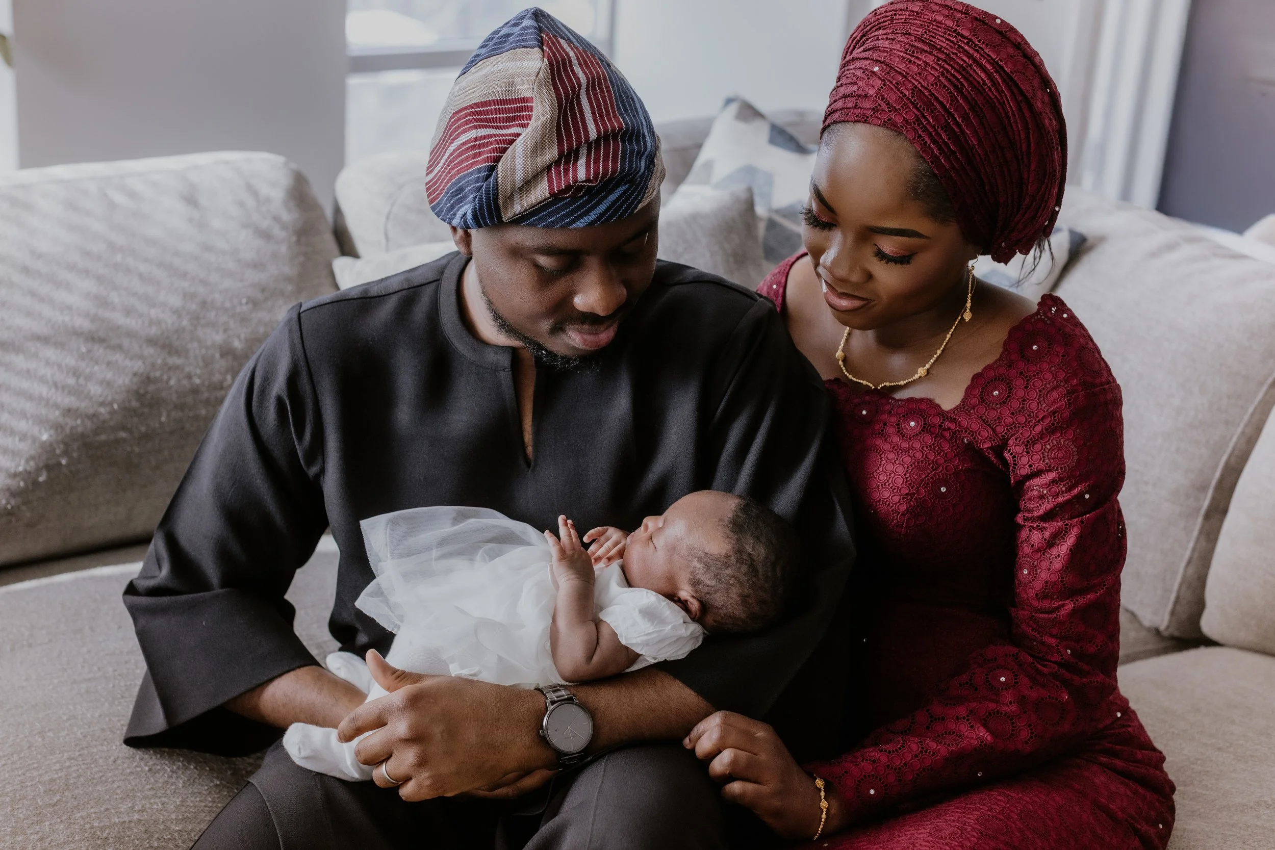 A man and woman sitting on a beige couch holding a newborn baby together. The man is wearing traditional African attire with a matching cap, and the woman is dressed in a burgundy lace dress with a matching headwrap. They are both looking at the baby