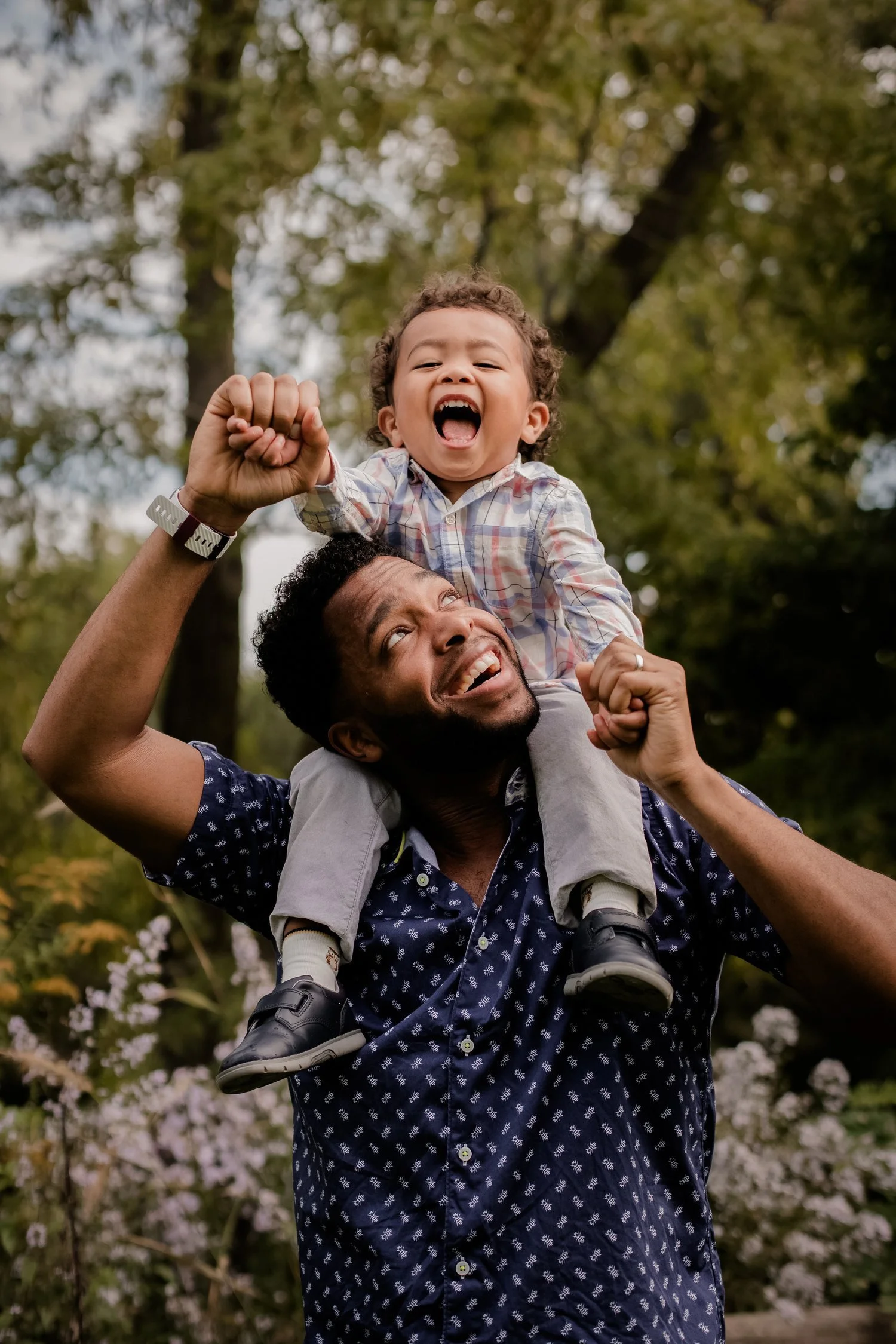 A joyful man carrying a young boy on his shoulders during an outdoor outing, with trees and flowers in the background.