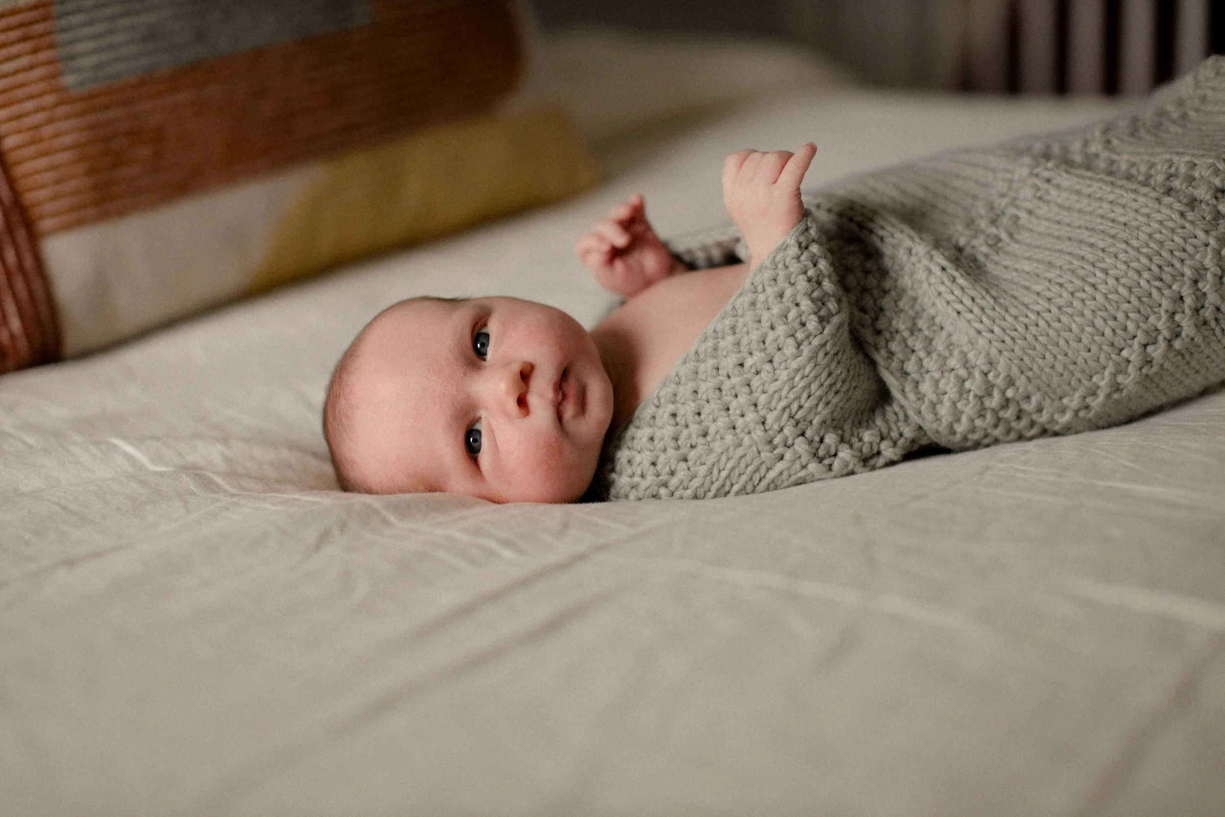 A baby lying on a bed, partially covered with a knitted blanket, looking at the camera. Lifestyle newborn session shot in Chicago IL.