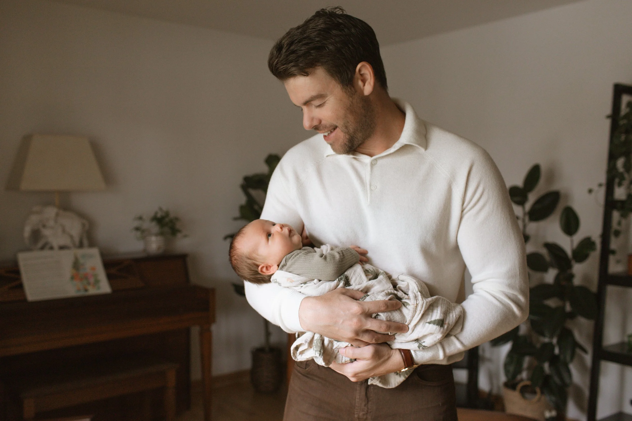 A man holding a newborn baby indoors, smiling while looking at the baby. Newborn photography in Chicago IL