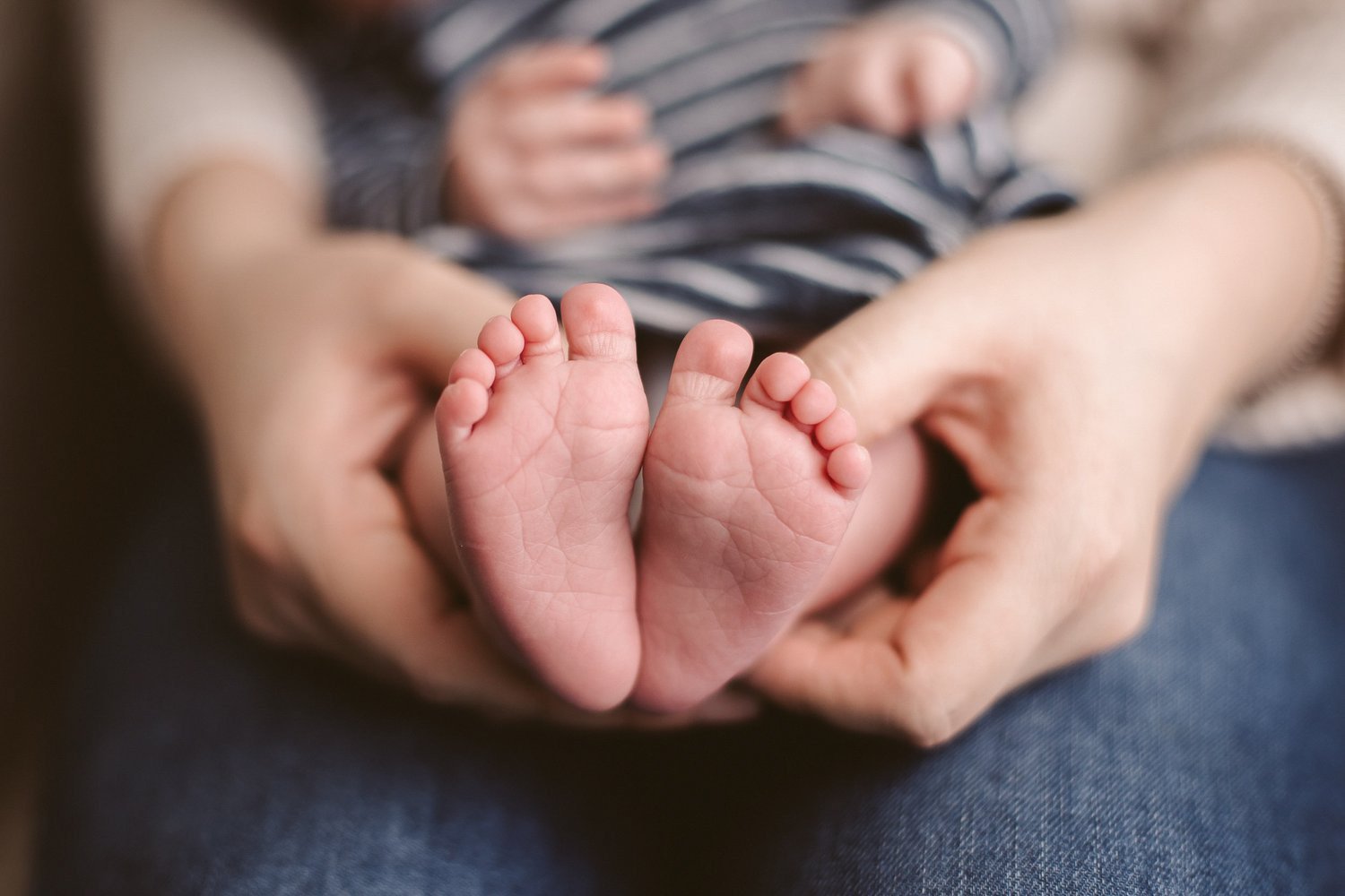Close-up of a baby's tiny feet being gently held by an adult's hands, with the baby wearing striped clothing.