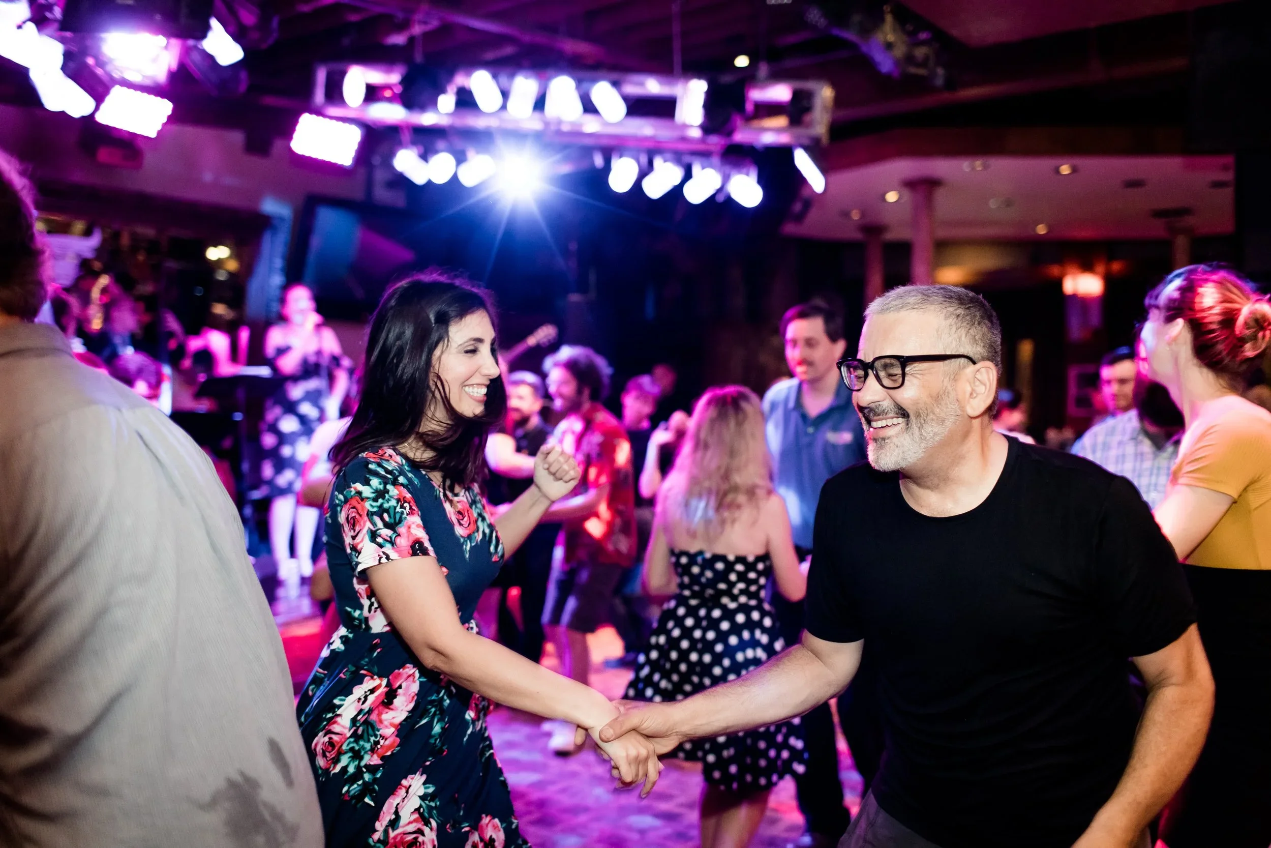 People dancing and socializing at a lively indoor party with colorful lighting, a live band performing on stage in the background, and couples smiling and shaking hands on the dance floor.  Dance, Jazz Photography Chicago IL, Erin Drewitz Photography