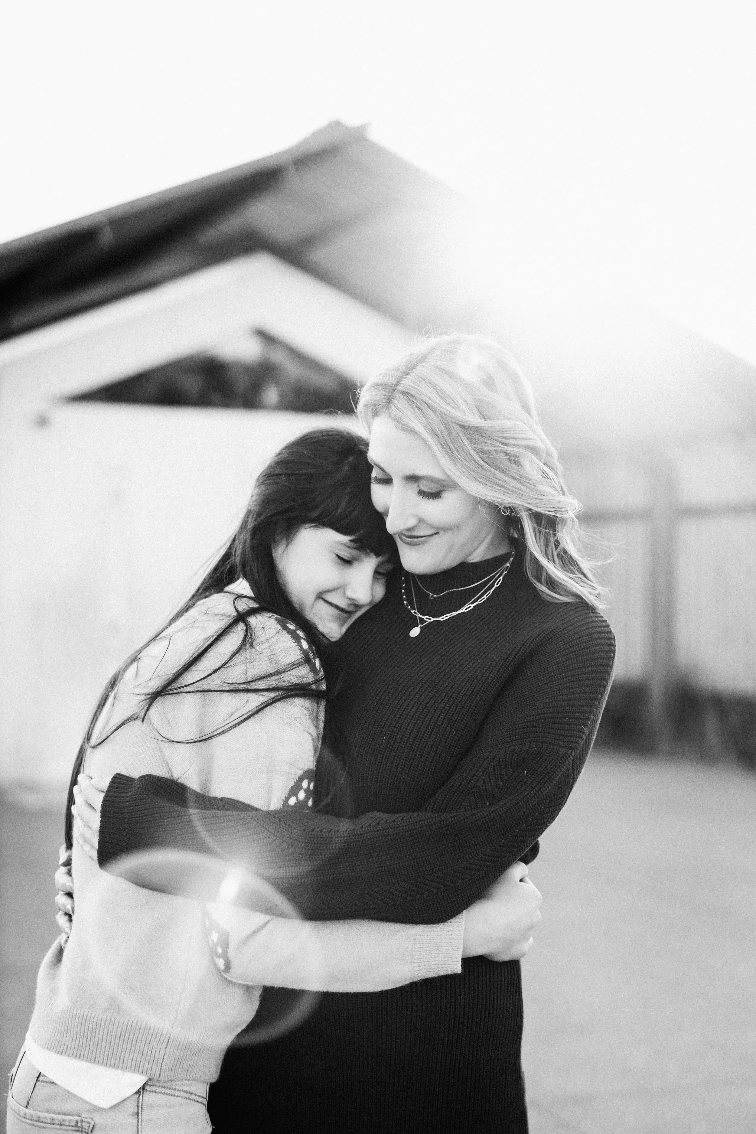 A girl with dark hair and a woman with blonde hair embrace outdoors, both smiling with closed eyes.