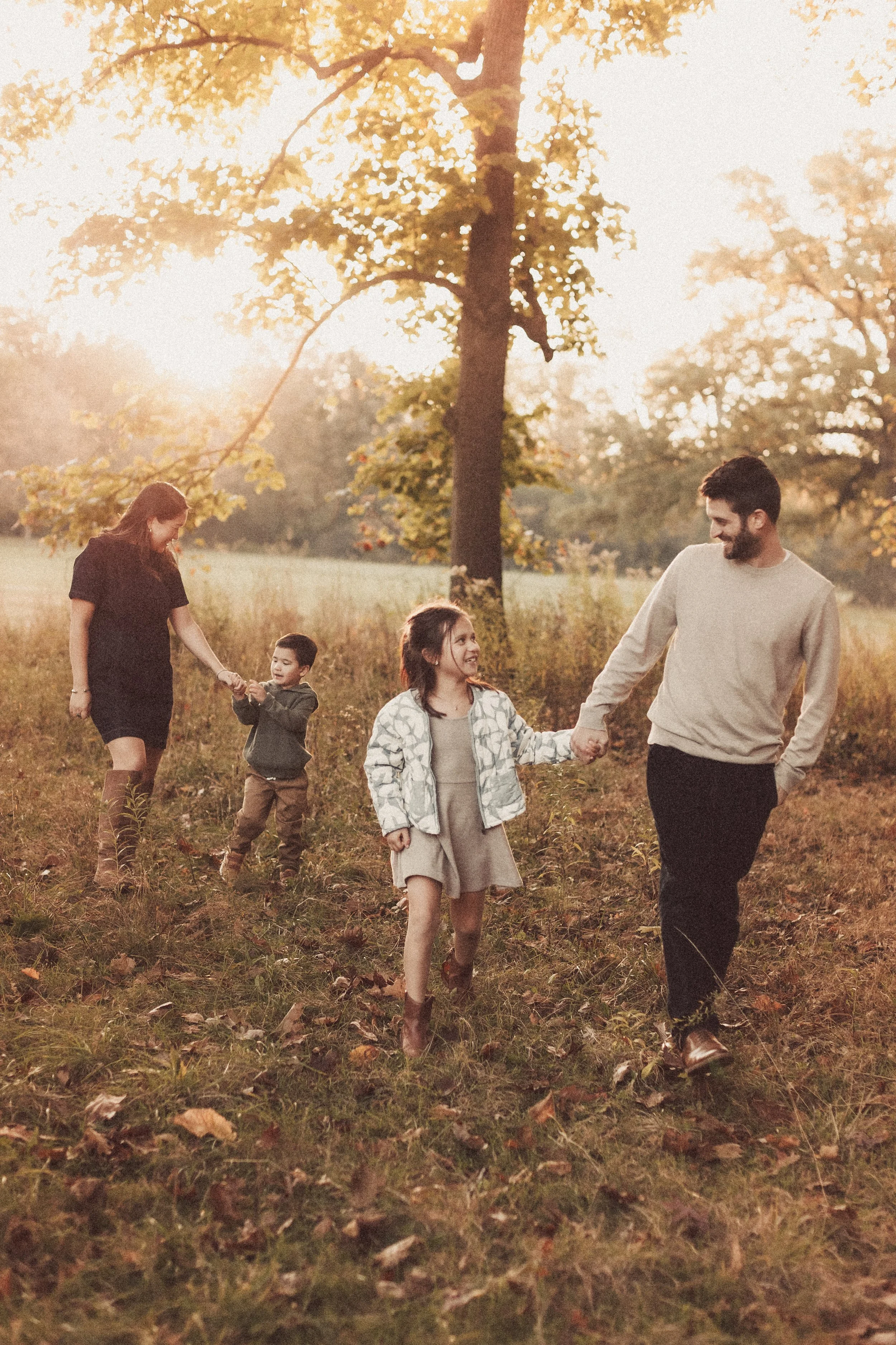 A family of four walking and holding hands in a park during autumn. The sunlight filters through the trees, creating a warm glow.  Natural documentary family photography shot in Harms Woods, Glenview IL. Family photographer based in Evanston.