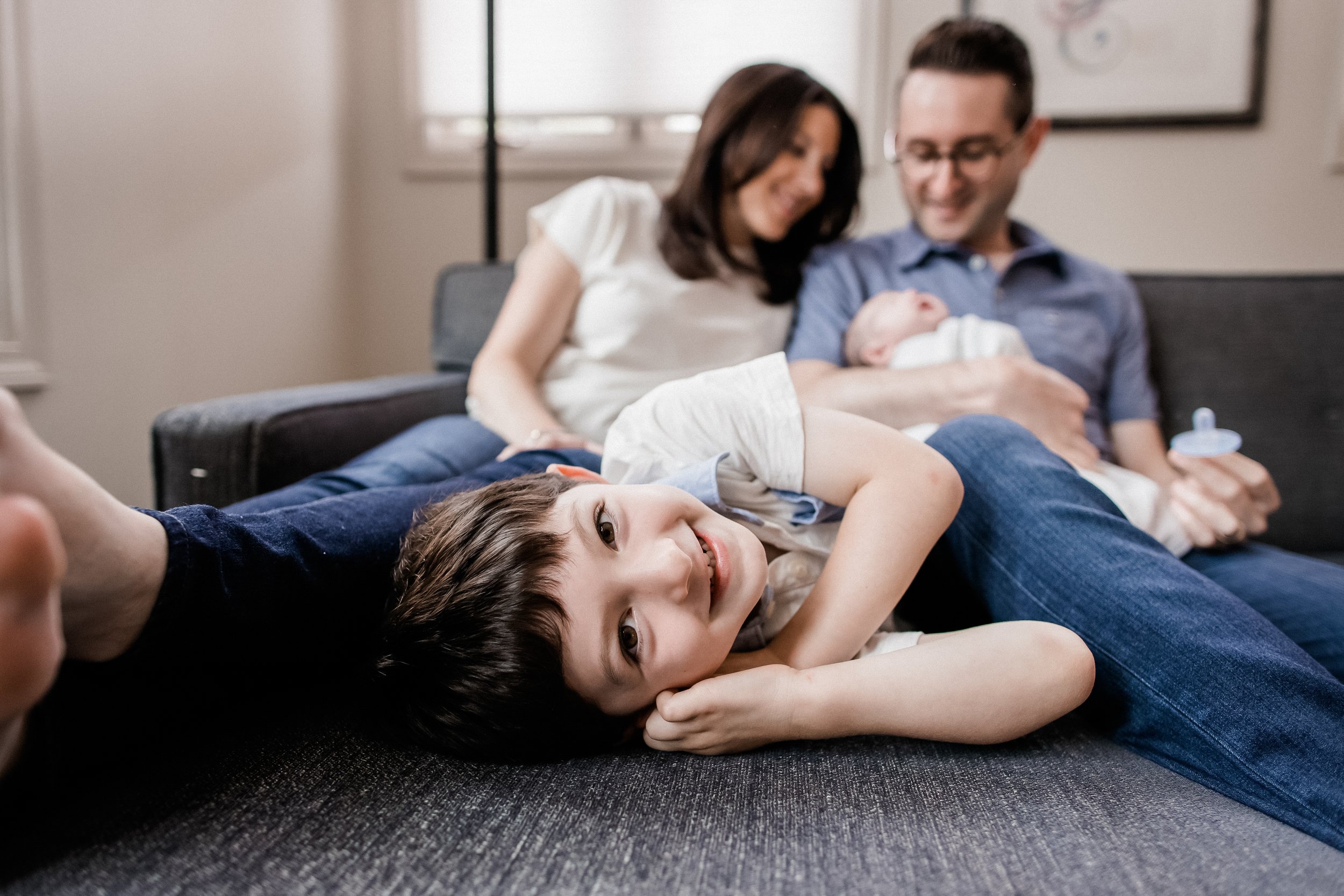 A family of four relaxing on a sofa in a living room. A young boy with dark hair is lying on the sofa with his head resting on a sibling's arm, smiling at the camera. In the background, a woman and a man are sitting close together, holding a newborn 