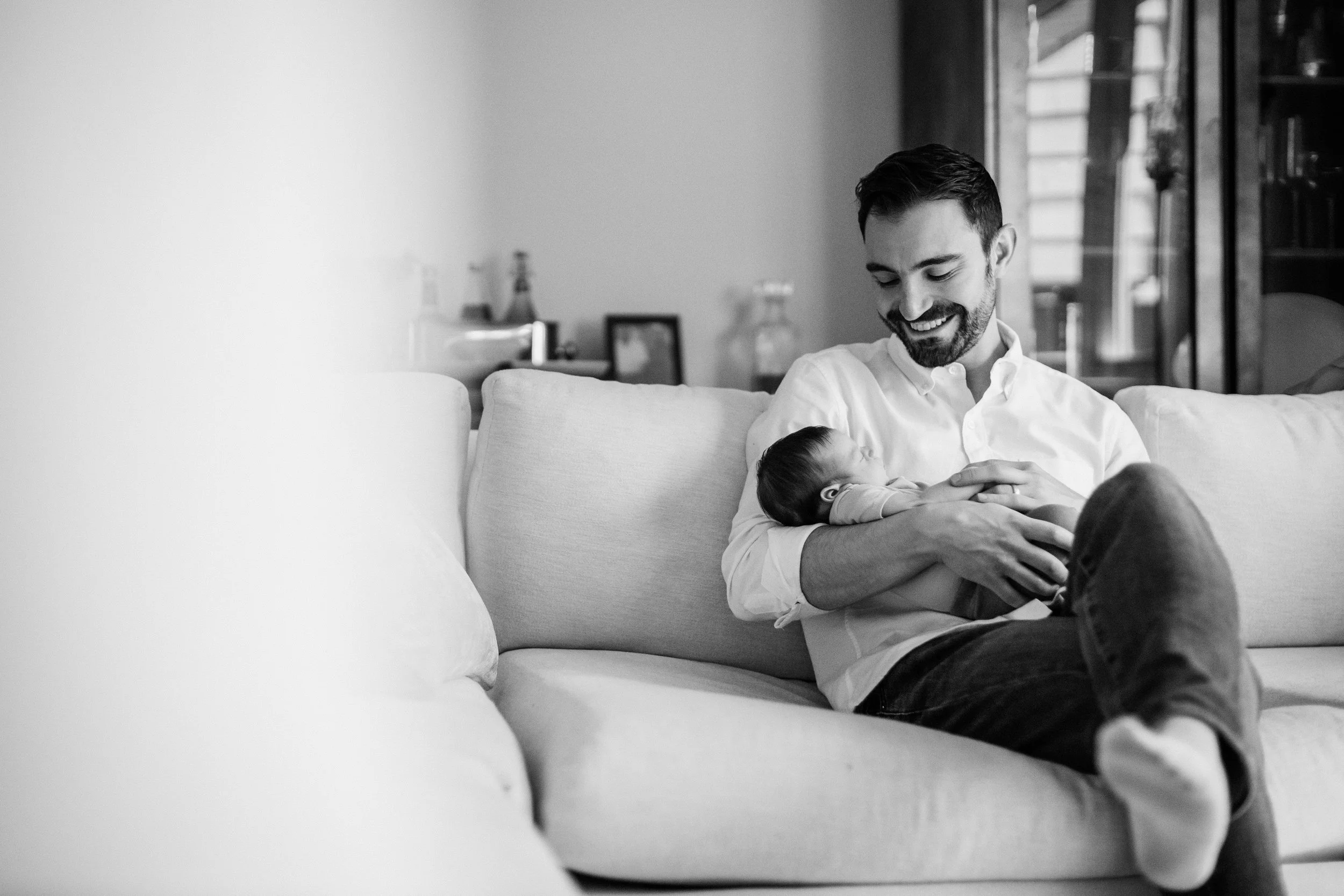 A man sitting on a sofa holding and looking at a newborn baby, both smiling, in a well-lit living room. Lifestyle newborn session shot in Skokie IL.