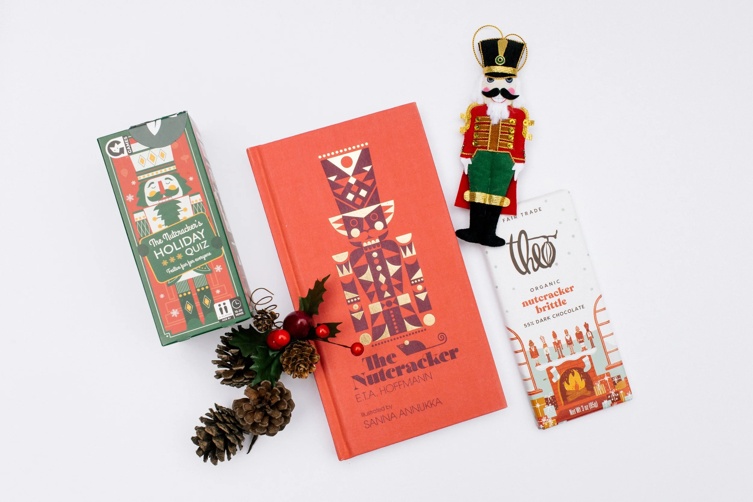 A Christmas-themed flat lay includes a holiday quiz box, a book titled "The Nutcracker" by E.T.A. Hoffmann, a decorative nutcracker figure, a chocolate bar, and a small pinecone and holly leaf with berries. Product Photography Shoot Evanston, IL.