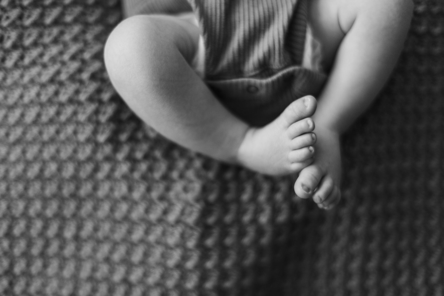 Black and white photo of a child sitting cross-legged on a patterned carpet, wearing a sleeveless top, with arms crossed over knees and hands clasped. Lifestyle newborn session shot in Evanston IL.
