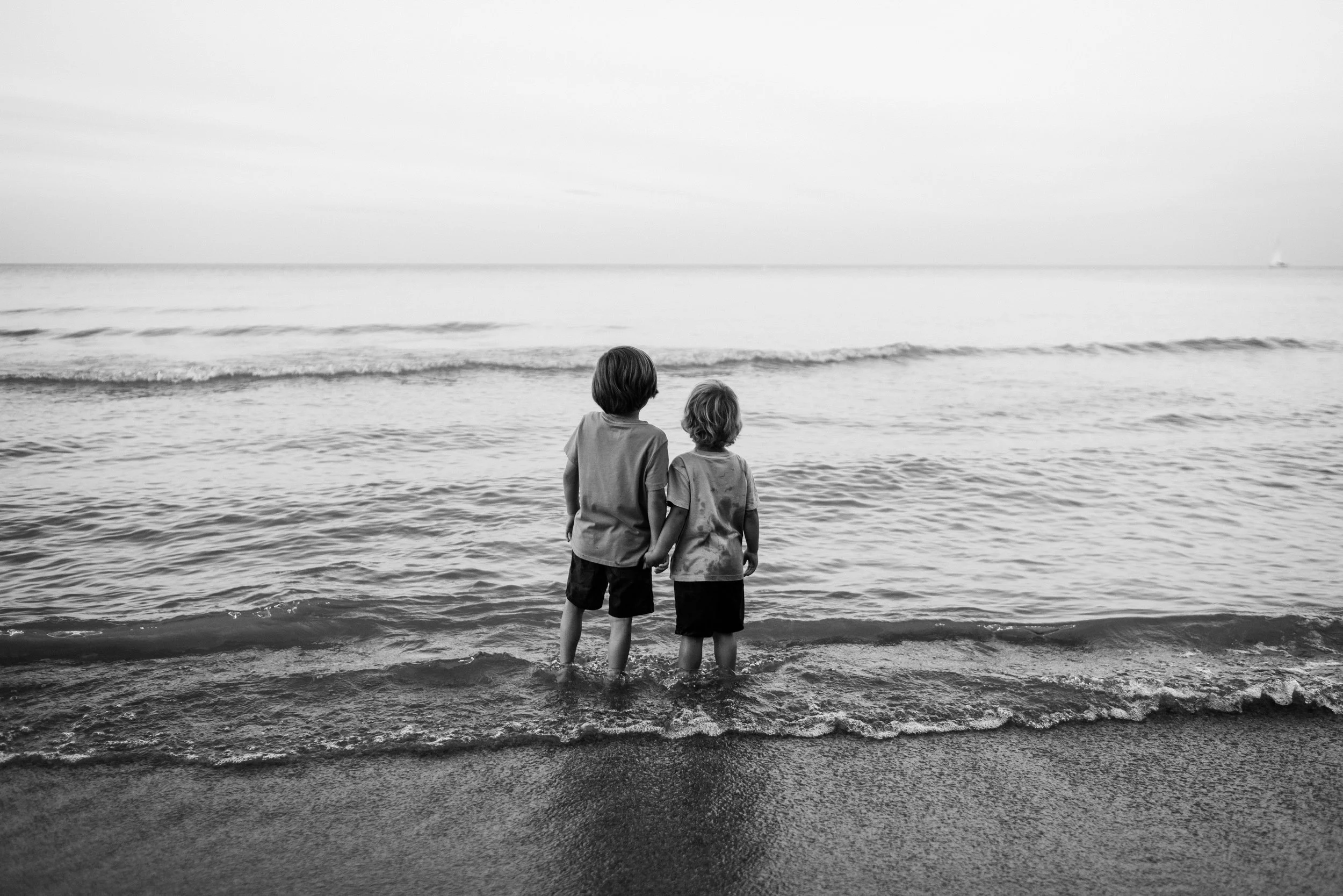 Two children holding hands, standing in shallow ocean water facing the horizon, in black and white.