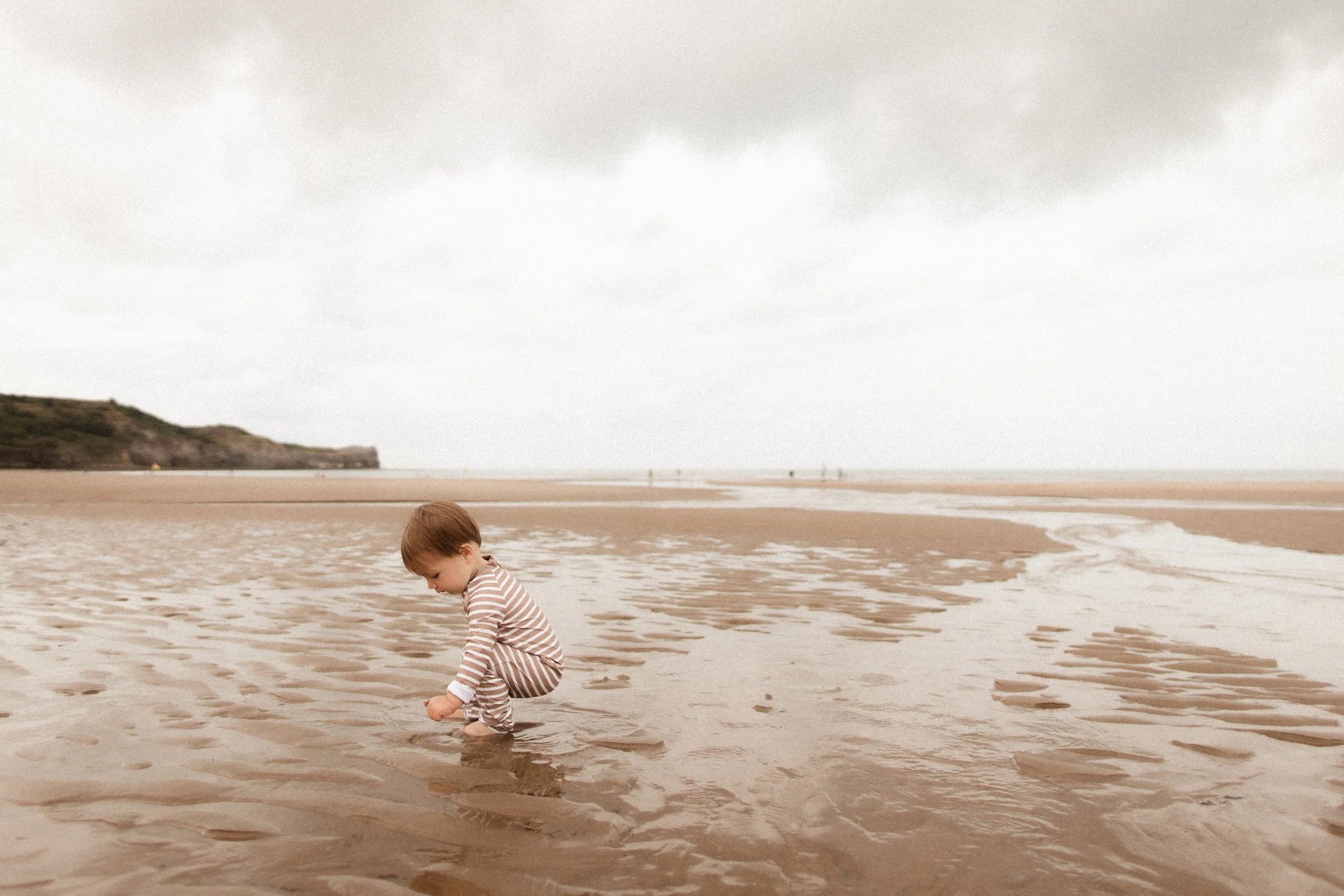 A young child in striped pajamas is crouched down in wet sand by the edge of the ocean, playing with the water and sand on a cloudy day.