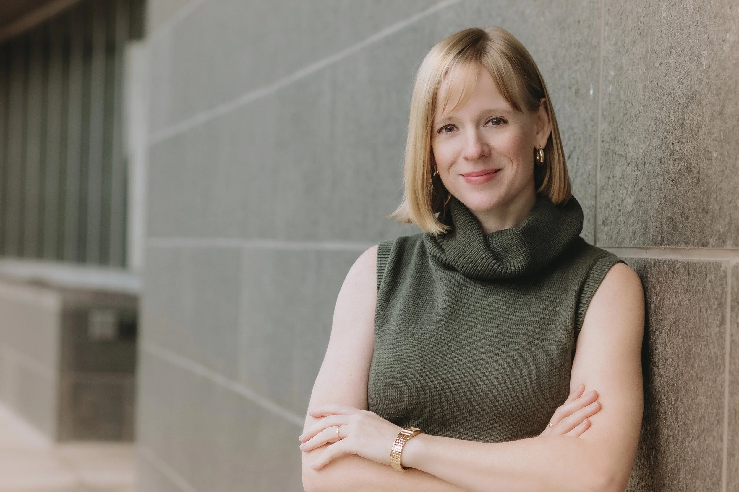 A woman with shoulder-length blonde hair, wearing a sleeveless dark green turtleneck sweater, gold hoop earrings, and a gold watch, leaning against a gray tiled wall and smiling at the camera.