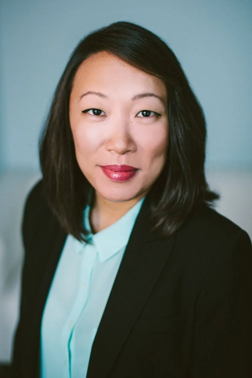 Professional headshot of a woman with black hair, wearing a white shirt with a black blazer, smiling slightly against a blue background. Headshot photography session in Chicago, IL.