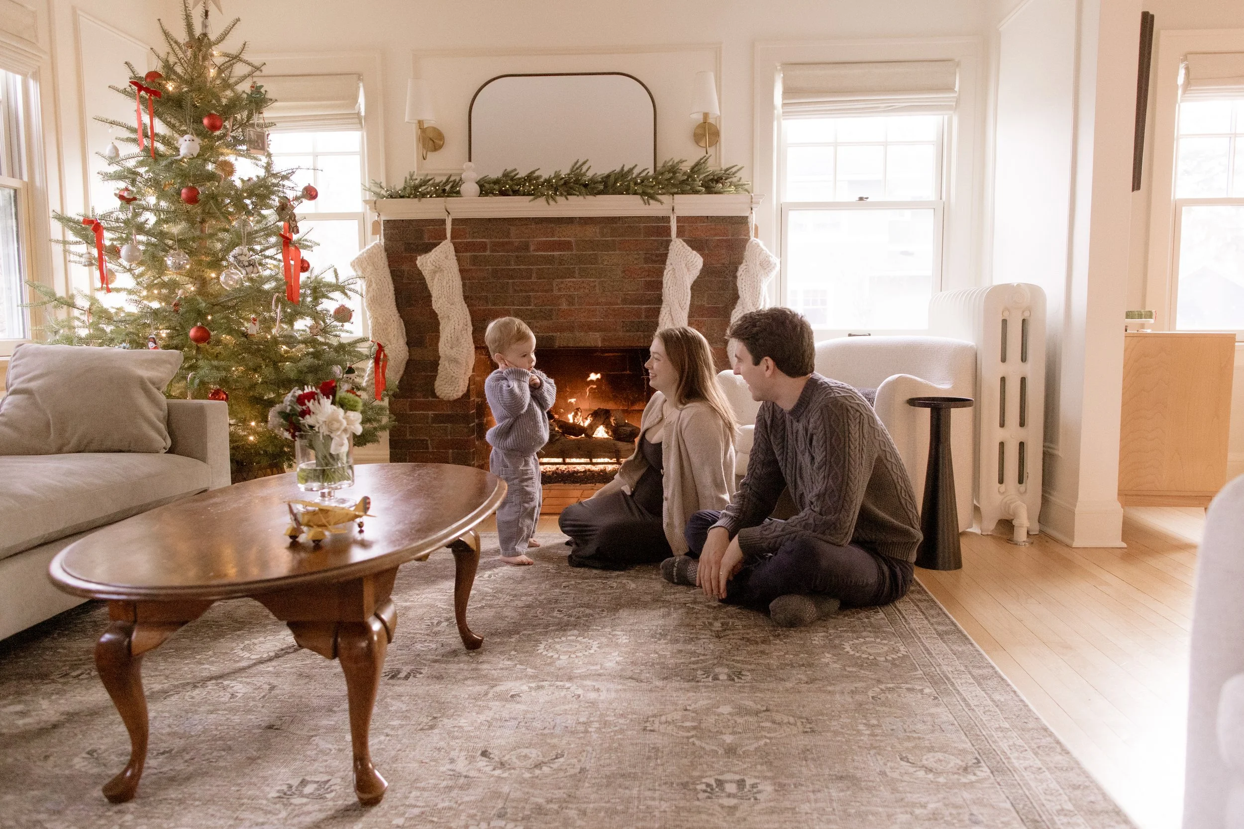 A family of three sitting by a lit fireplace in a cozy living room decorated for Christmas, with a decorated Christmas tree nearby. The child and mother are smiling at each other, and the father sits on the floor watching them.