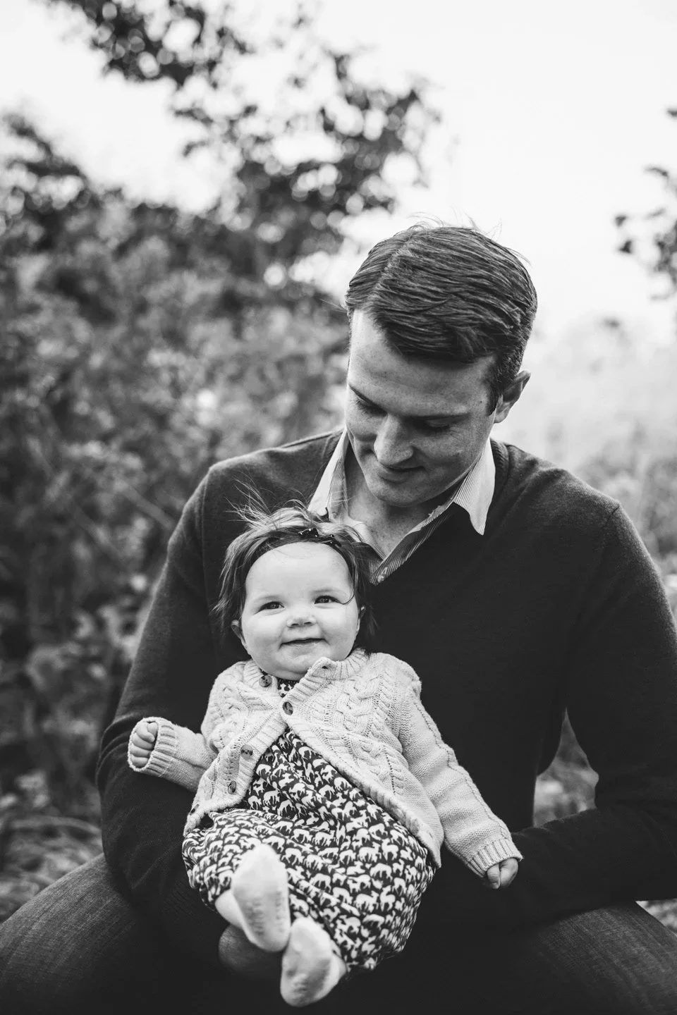 Black and white photo of a man holding a smiling baby girl outdoors with trees in the background. Film inspired family photography session in Chicago IL