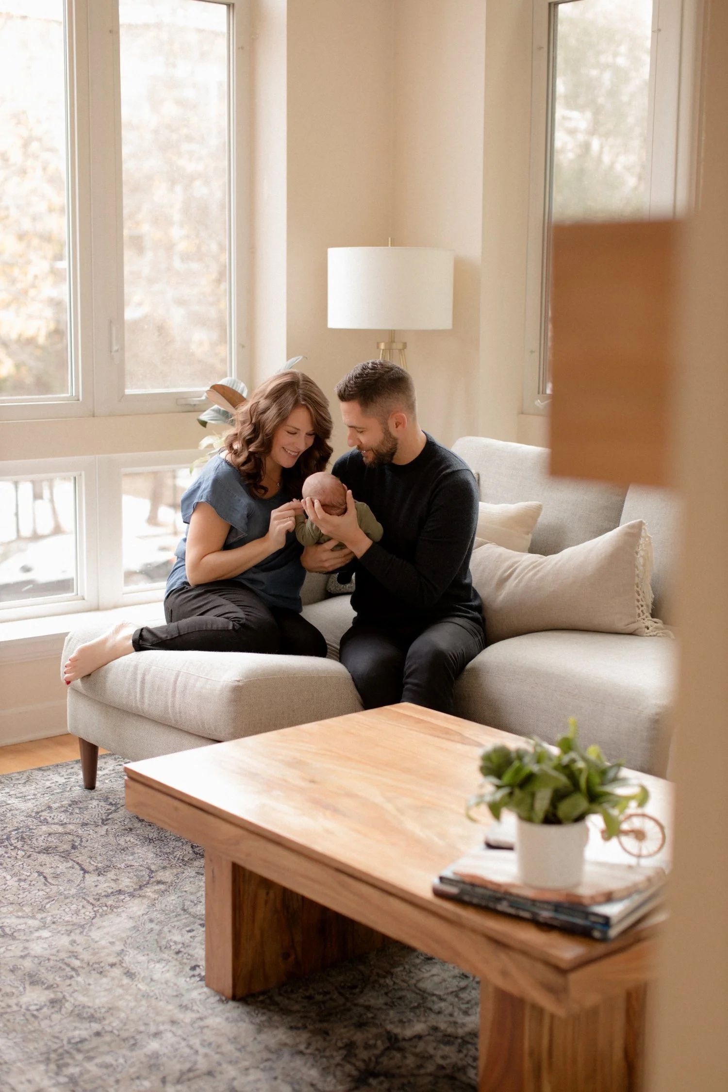 A happy couple sitting on a light-colored sofa in a well-lit living room, holding a newborn baby together and smiling at the baby. There is a large window behind them, a side table with a lamp, and a coffee table with a plant and magazines in the for