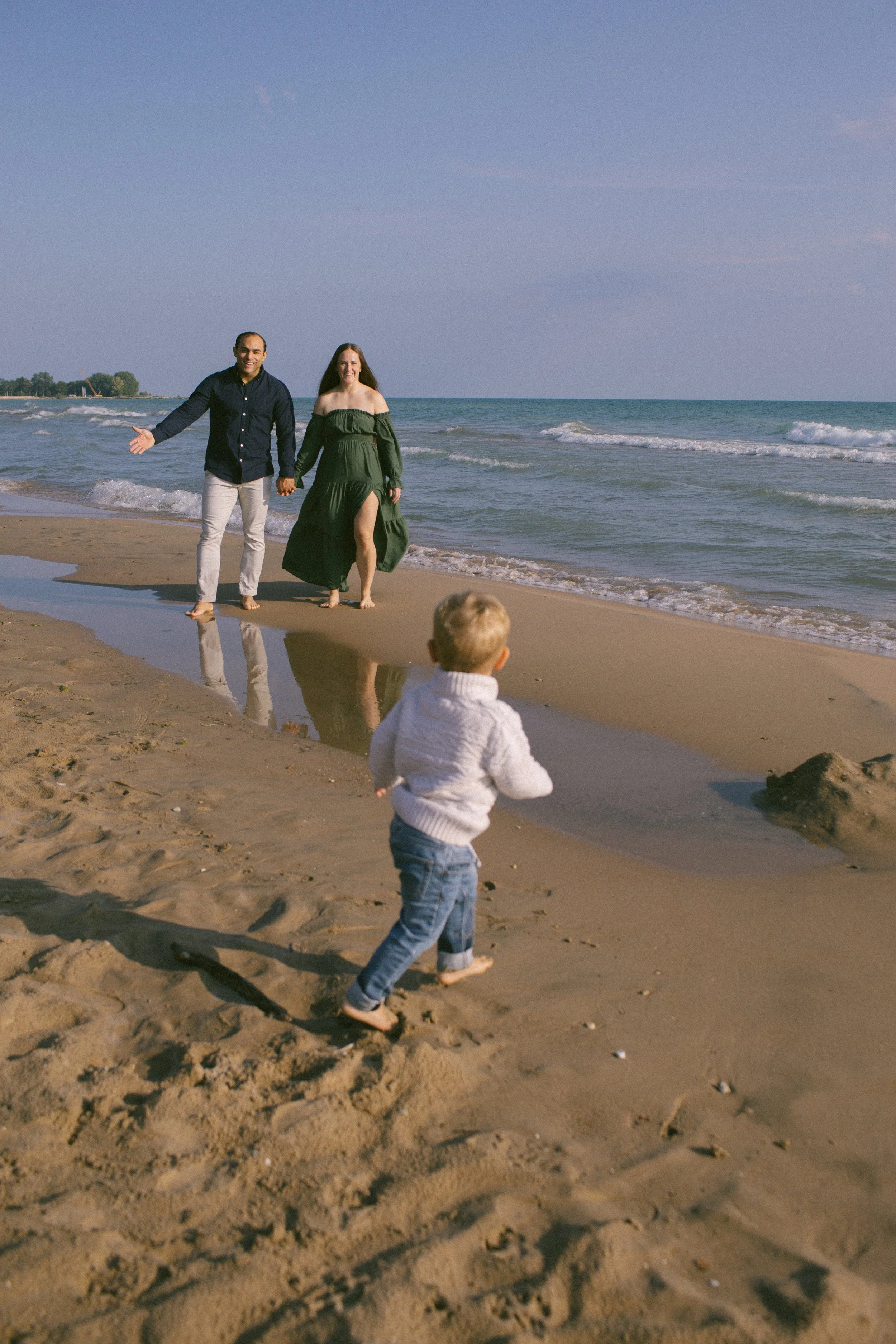 A family on a beach walk with a man and woman holding hands near the water, and a young boy running on the sand in the foreground. Shot at Evanston Lighthouse Beach Park, Grosse Point Lighthouse.  Lifestyle photography