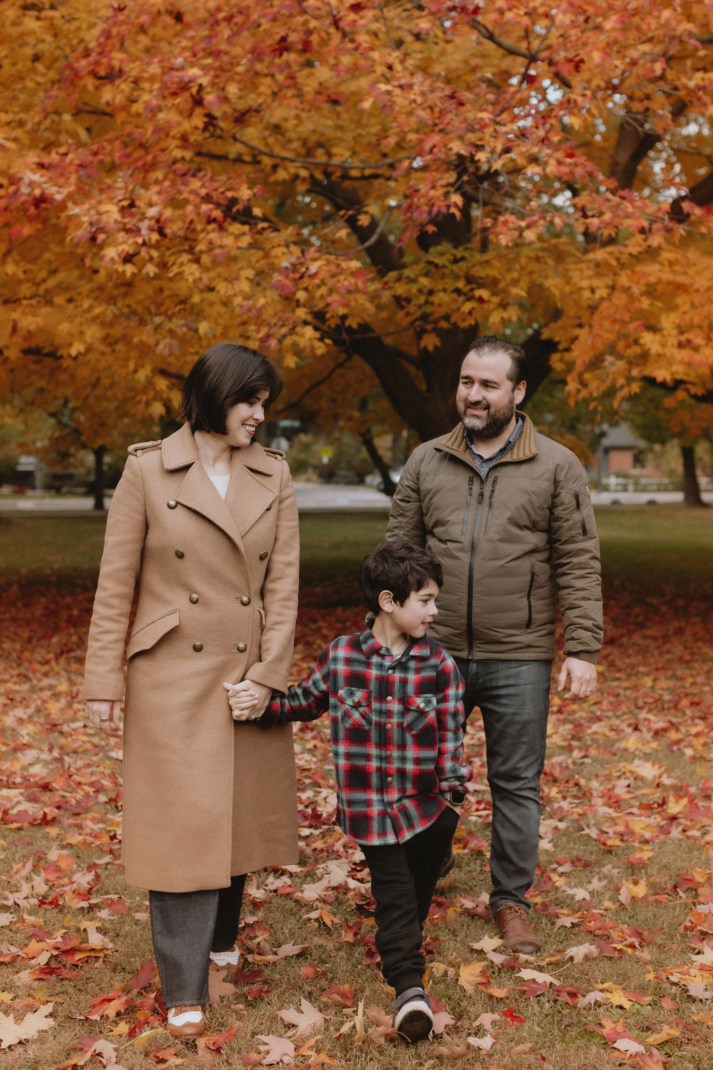 A family of three walking in a park during fall, with orange and red leaves on the ground and a large tree with vibrant autumn foliage in the background.