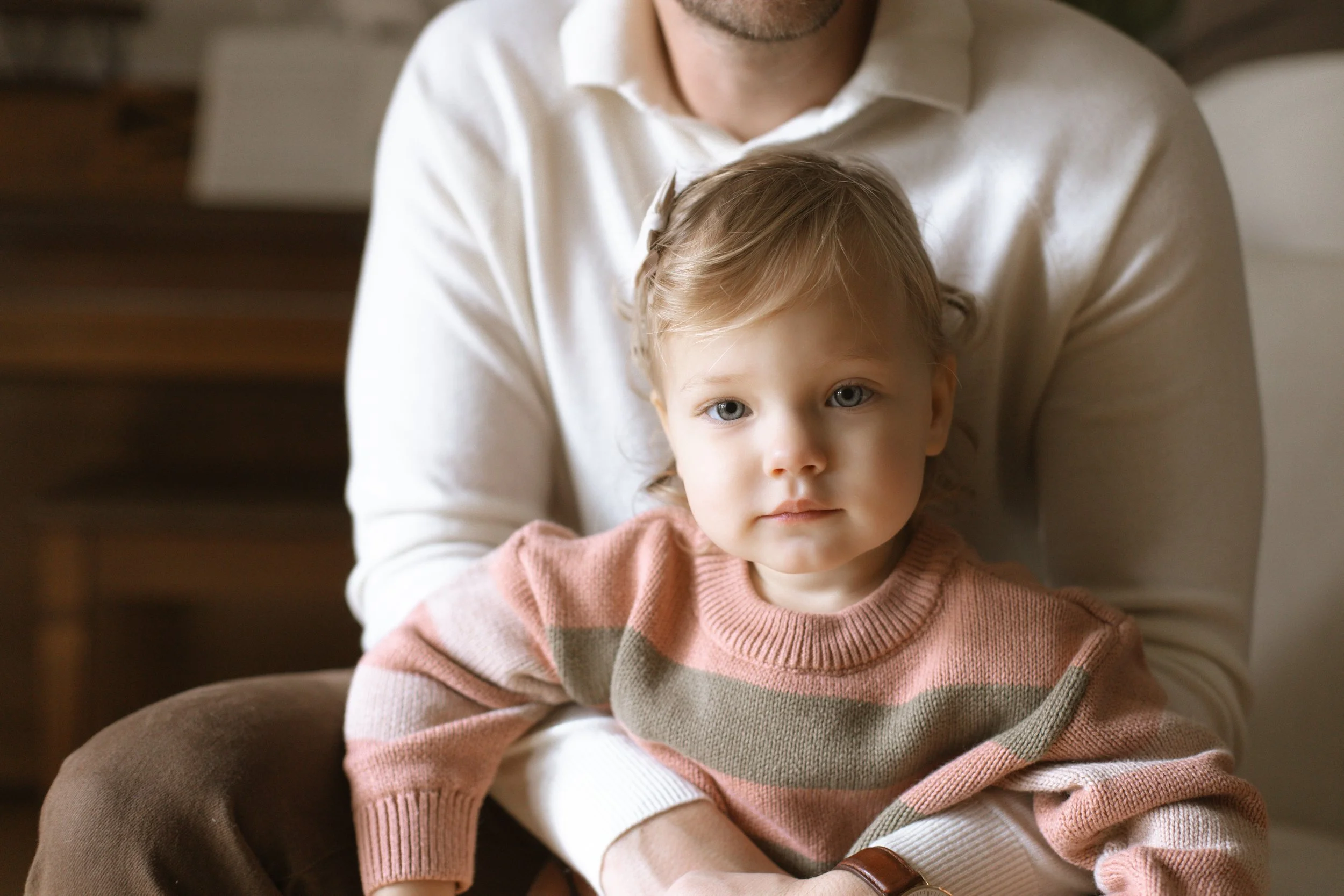 A young girl with blond hair and blue eyes sitting on an adult's lap, wearing a pink and gray striped sweater.